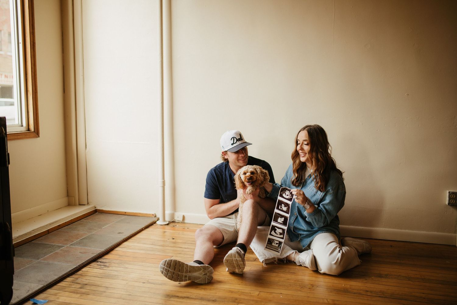 Couple sitting together on hardwood floor against beige wall.
