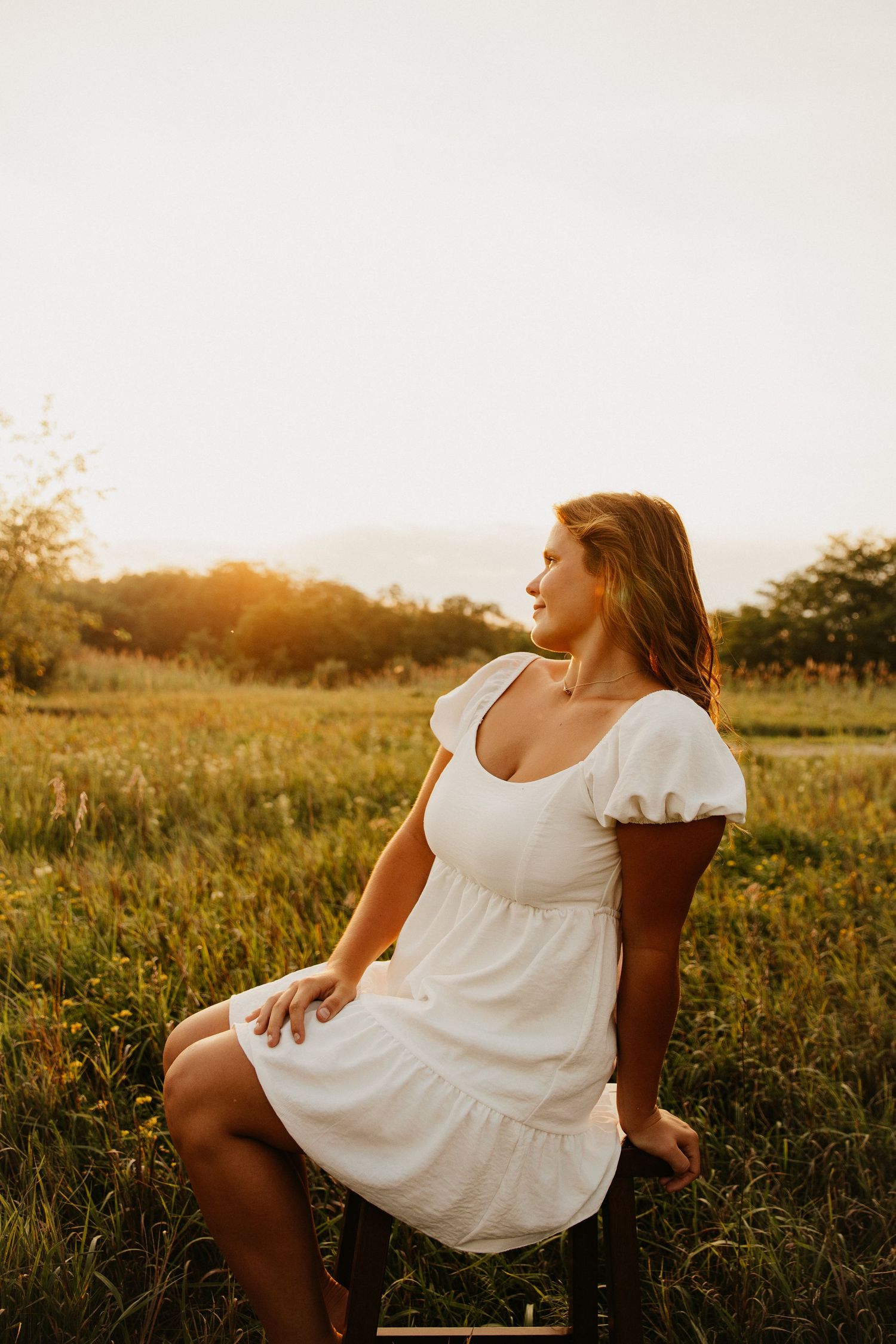 Person in flowing white dress sits in golden sunlit field during sunset.