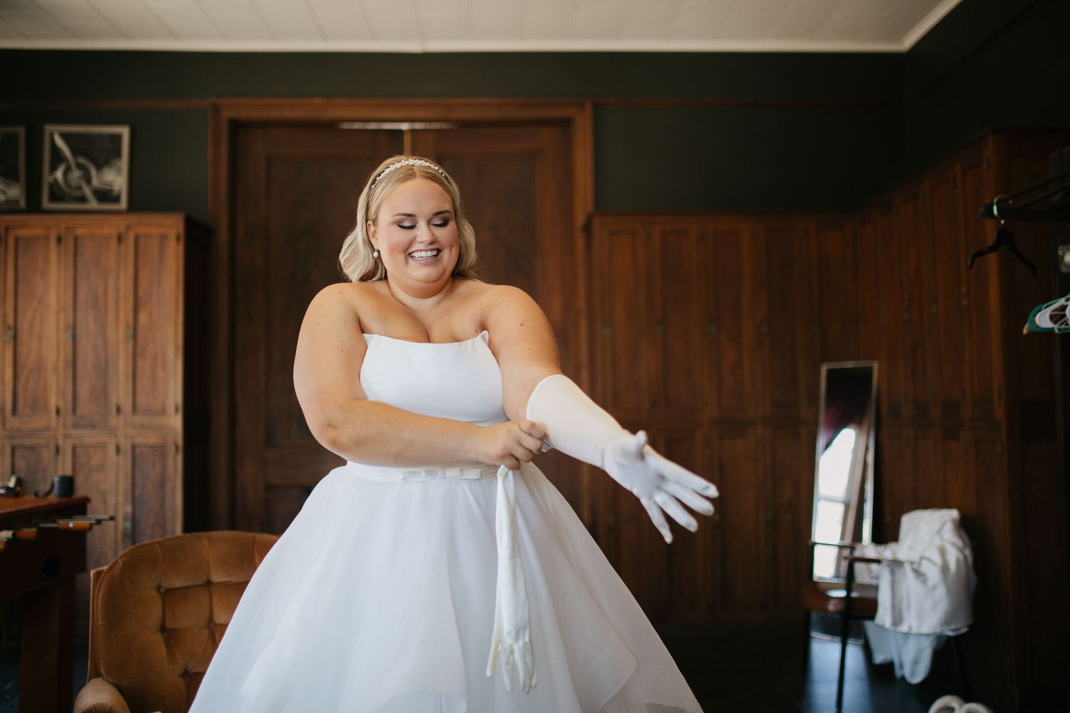 A person in a white wedding gown pulls on long gloves in a wood-paneled room.