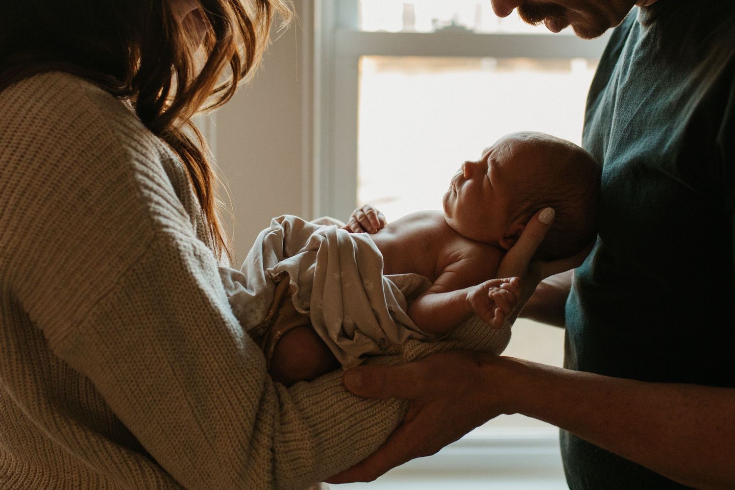 Tender moment of parents holding newborn baby near window in soft lighting.