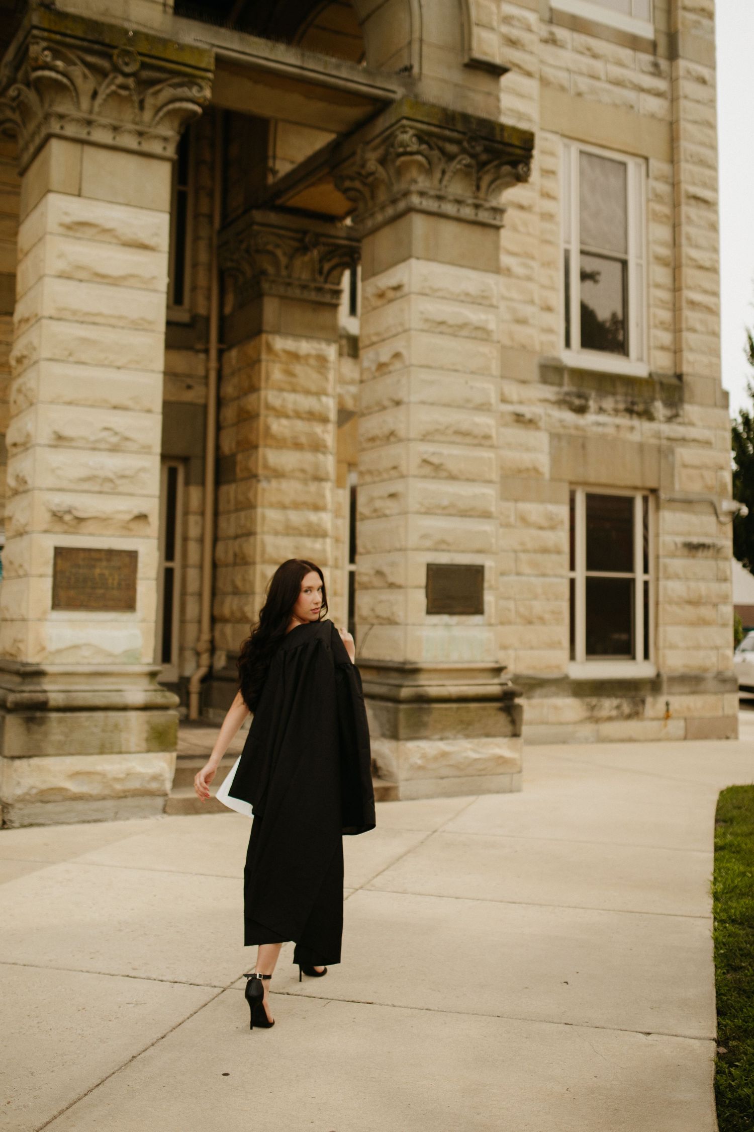 Figure in a black dress and boots walks along a stone building exterior in dramatic lighting.