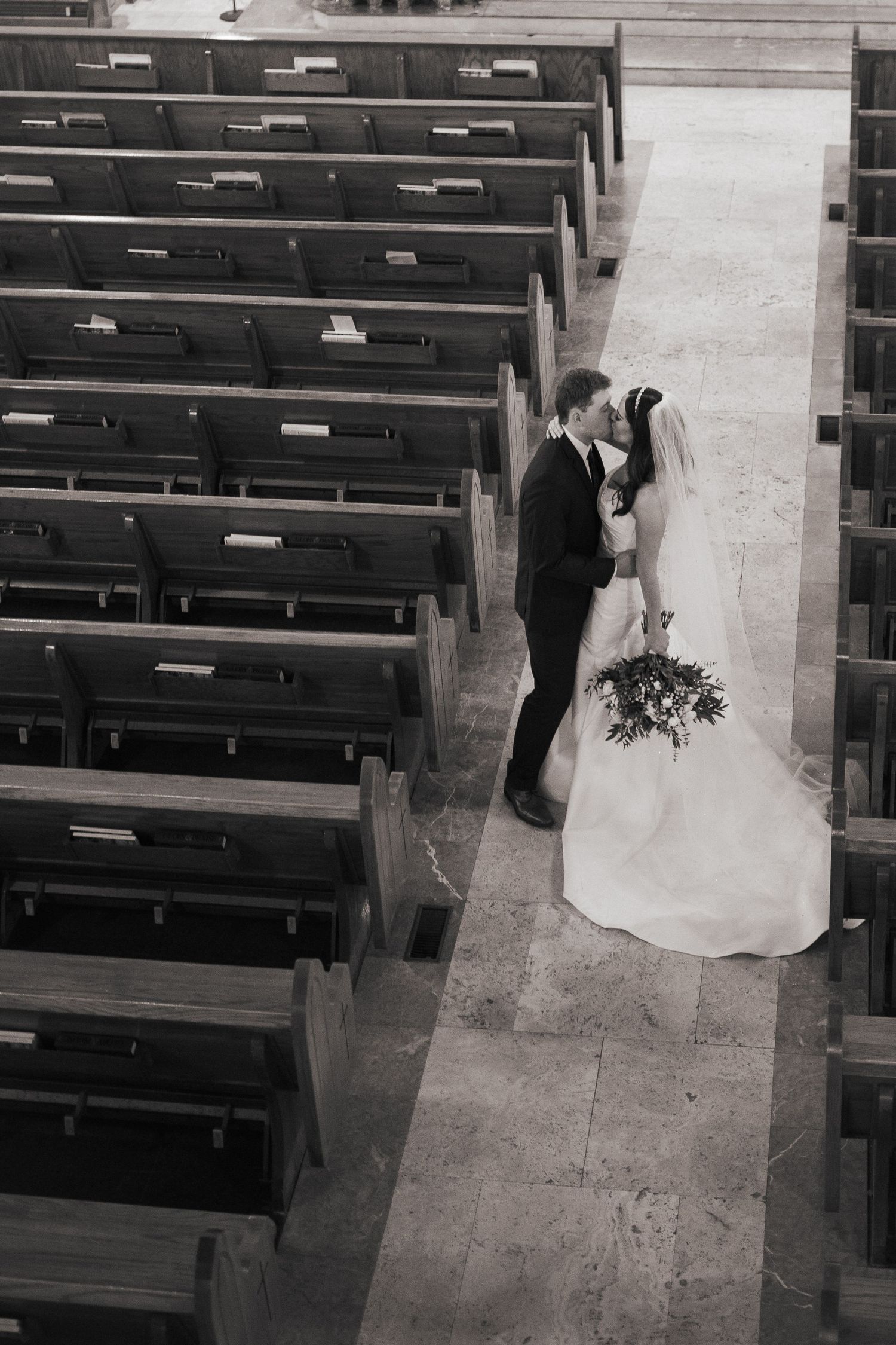 Black and white photo of a couple sharing an intimate moment in a church aisle surrounded by pews.