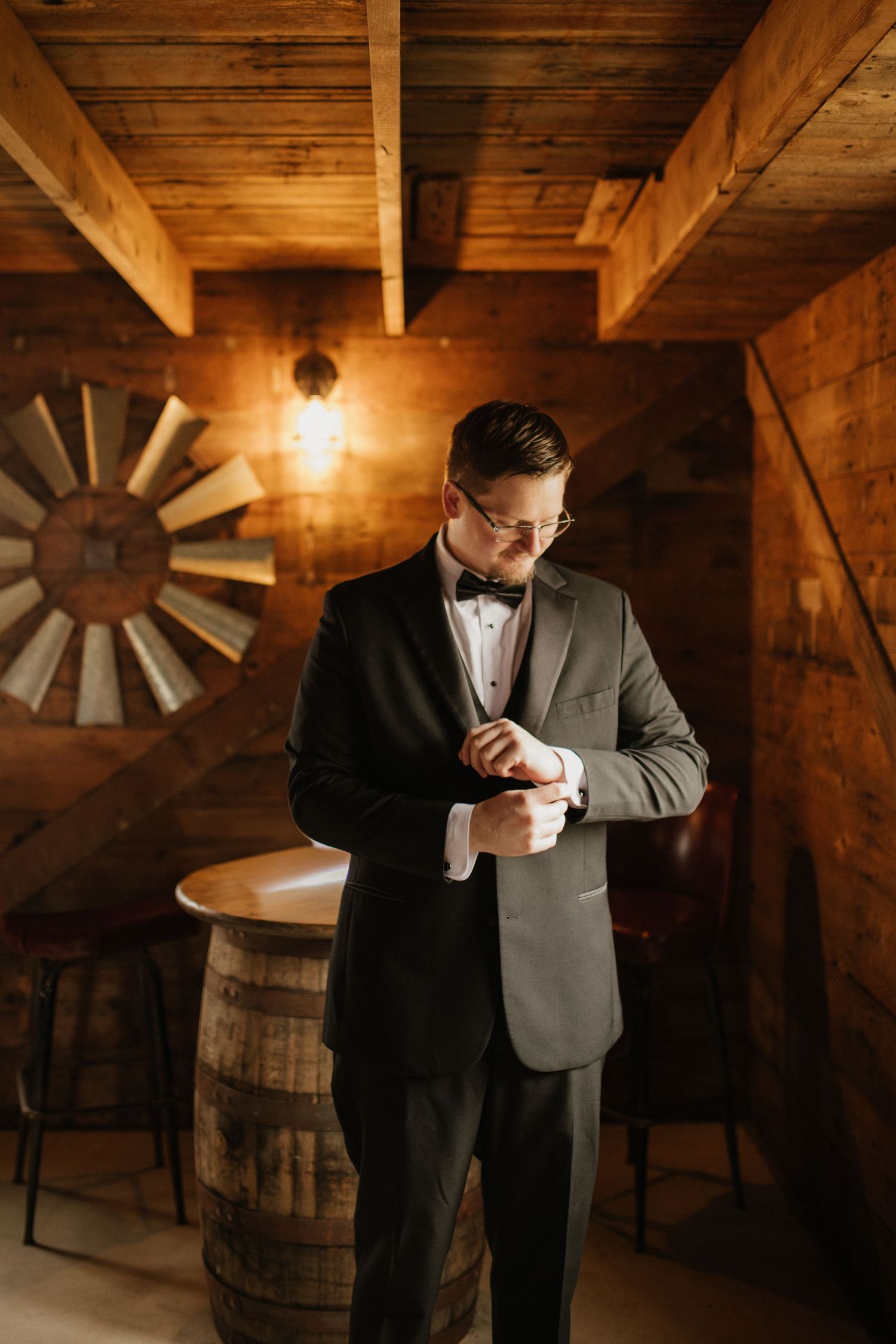 Groom adjusting suit in rustic wooden barn with ambient lighting.
