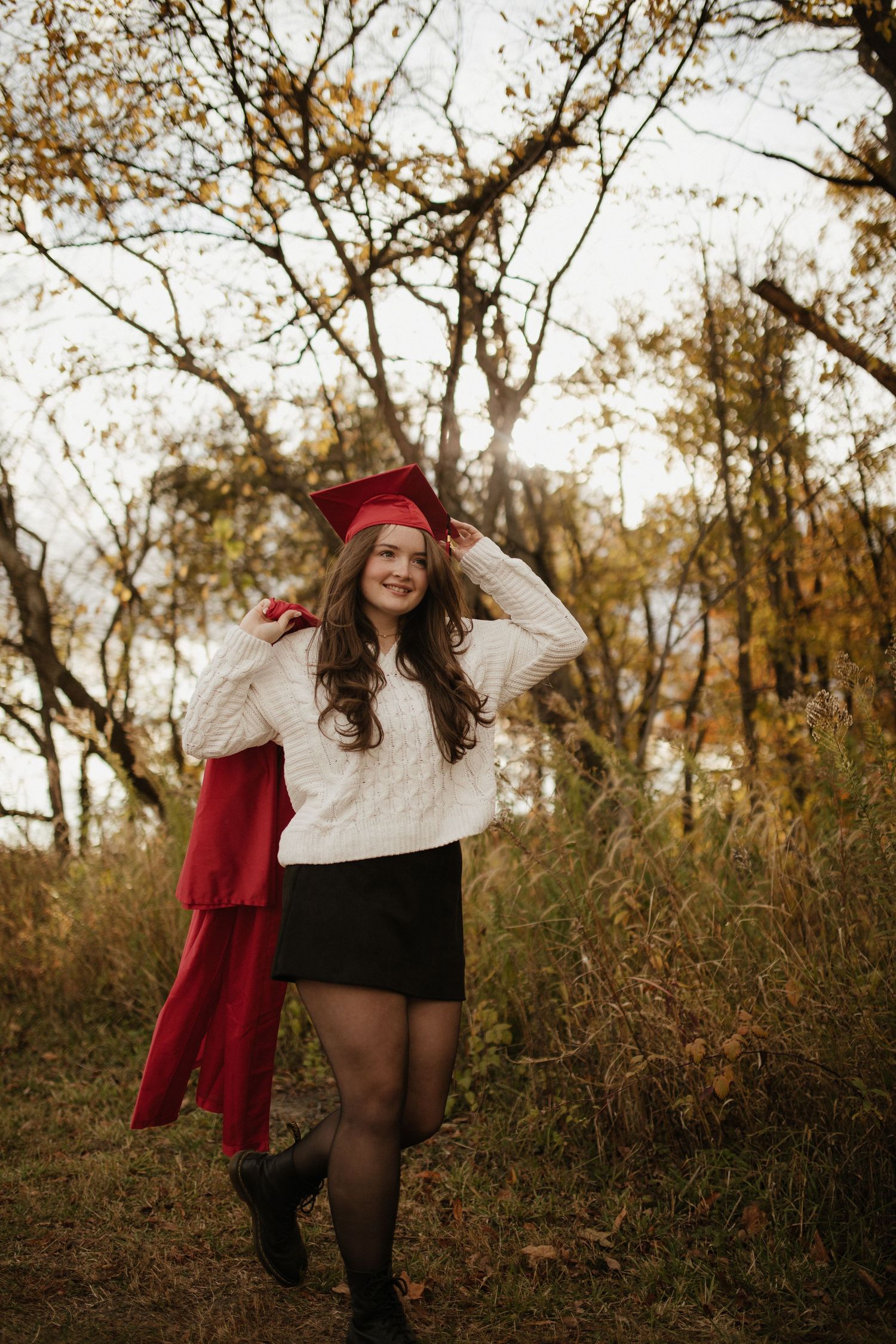 Someone in a red coat and white sweater playfully poses in a natural autumn setting.