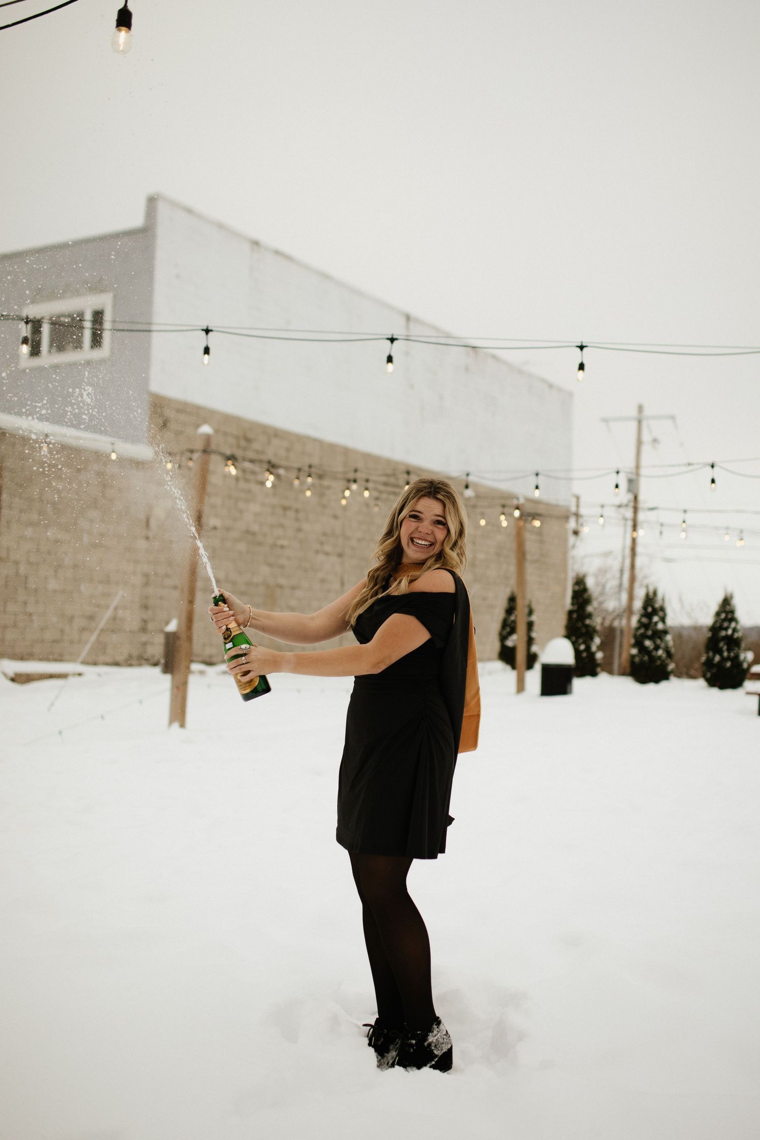 Graduate celebrating in snow with champagne bottle and string lights.