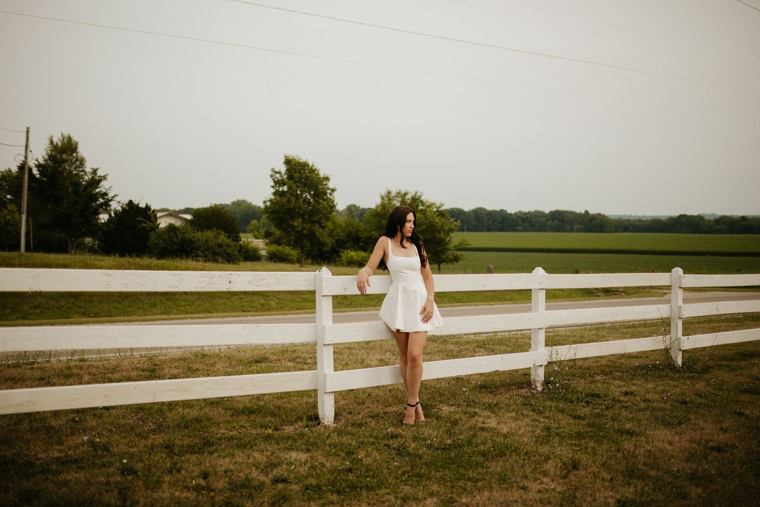 Person in white dress stands by rustic white fence in rural countryside setting.