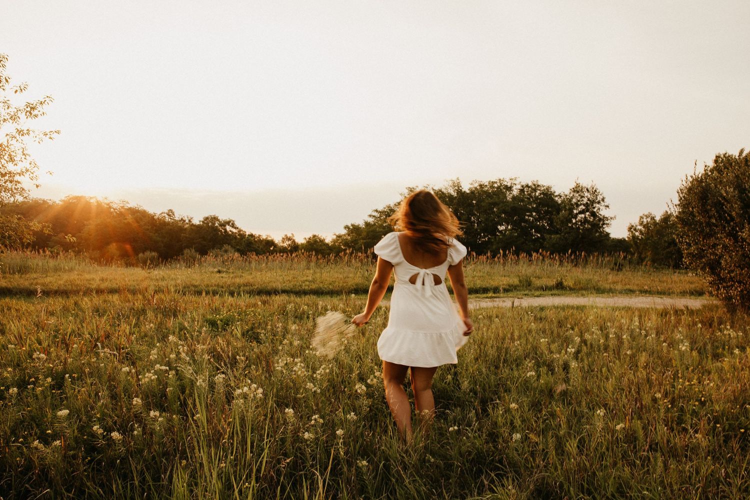 Figure in white dress stands among wildflowers in a meadow at sunset.