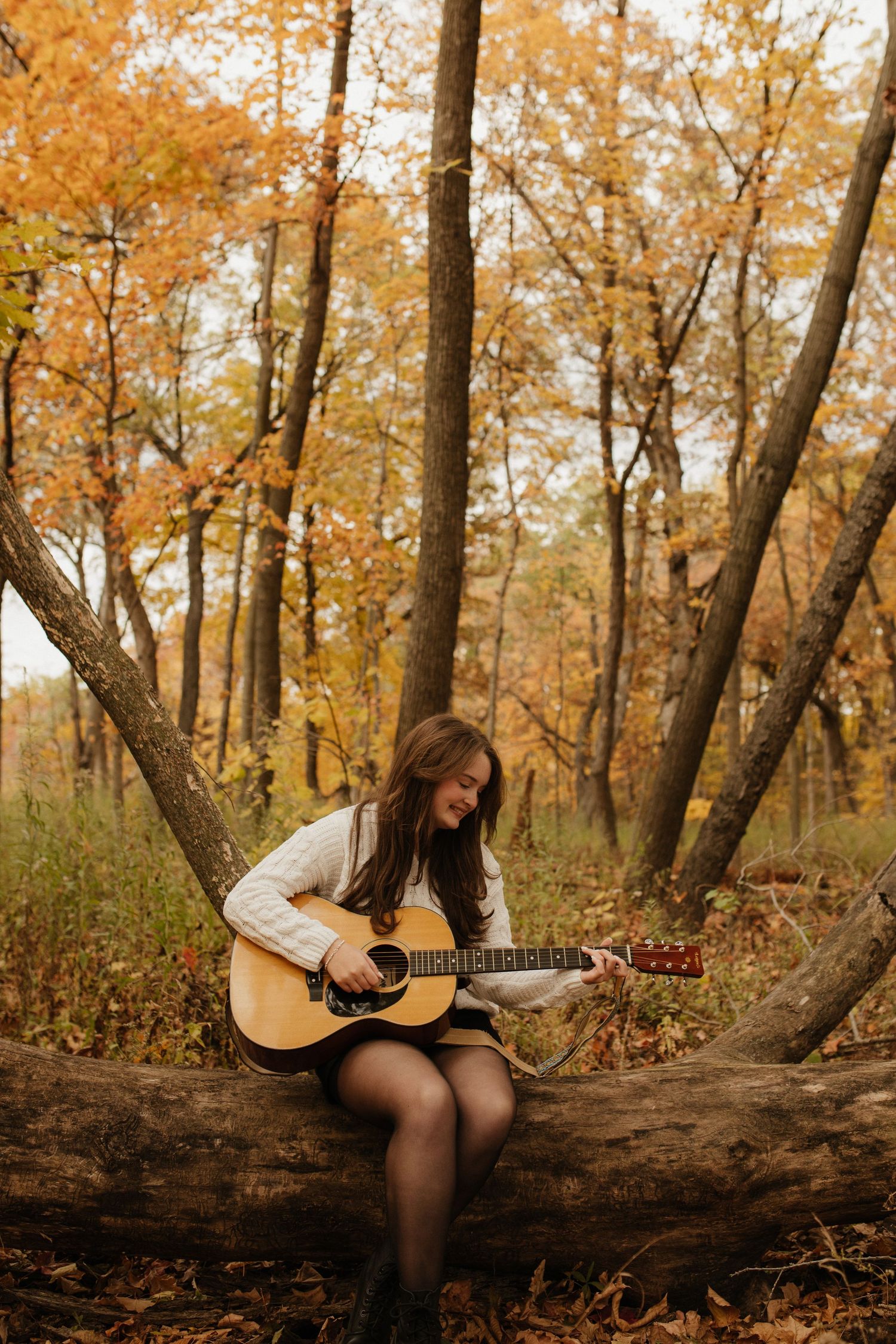A person sits on a fallen tree trunk playing acoustic guitar surrounded by golden autumn leaves.