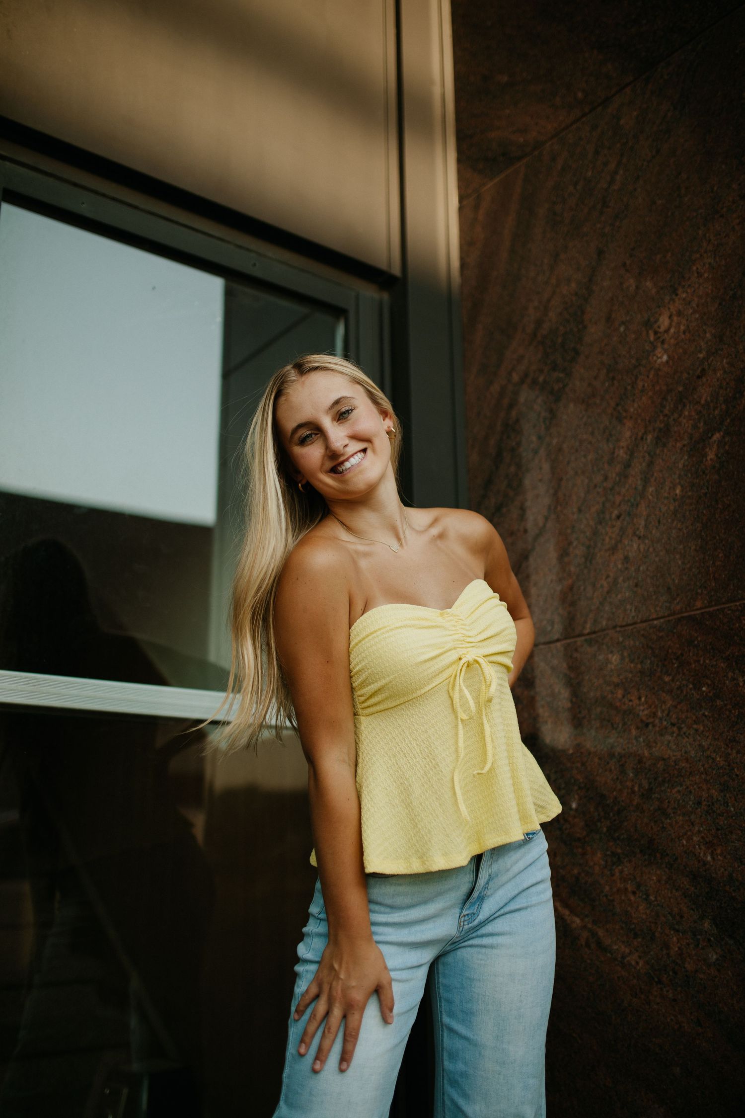 Person in yellow top and jeans poses against dark wooden wall.