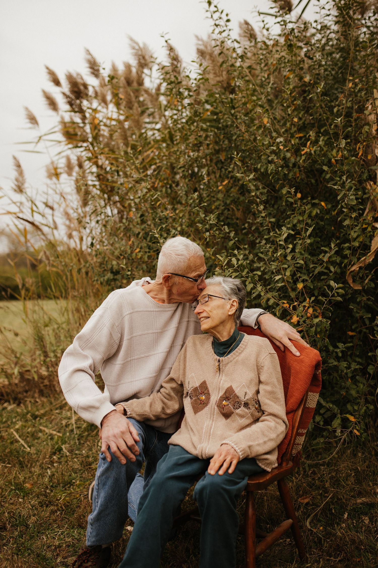 An elderly couple shares a tender moment while sitting on a wooden chair surrounded by tall grass.