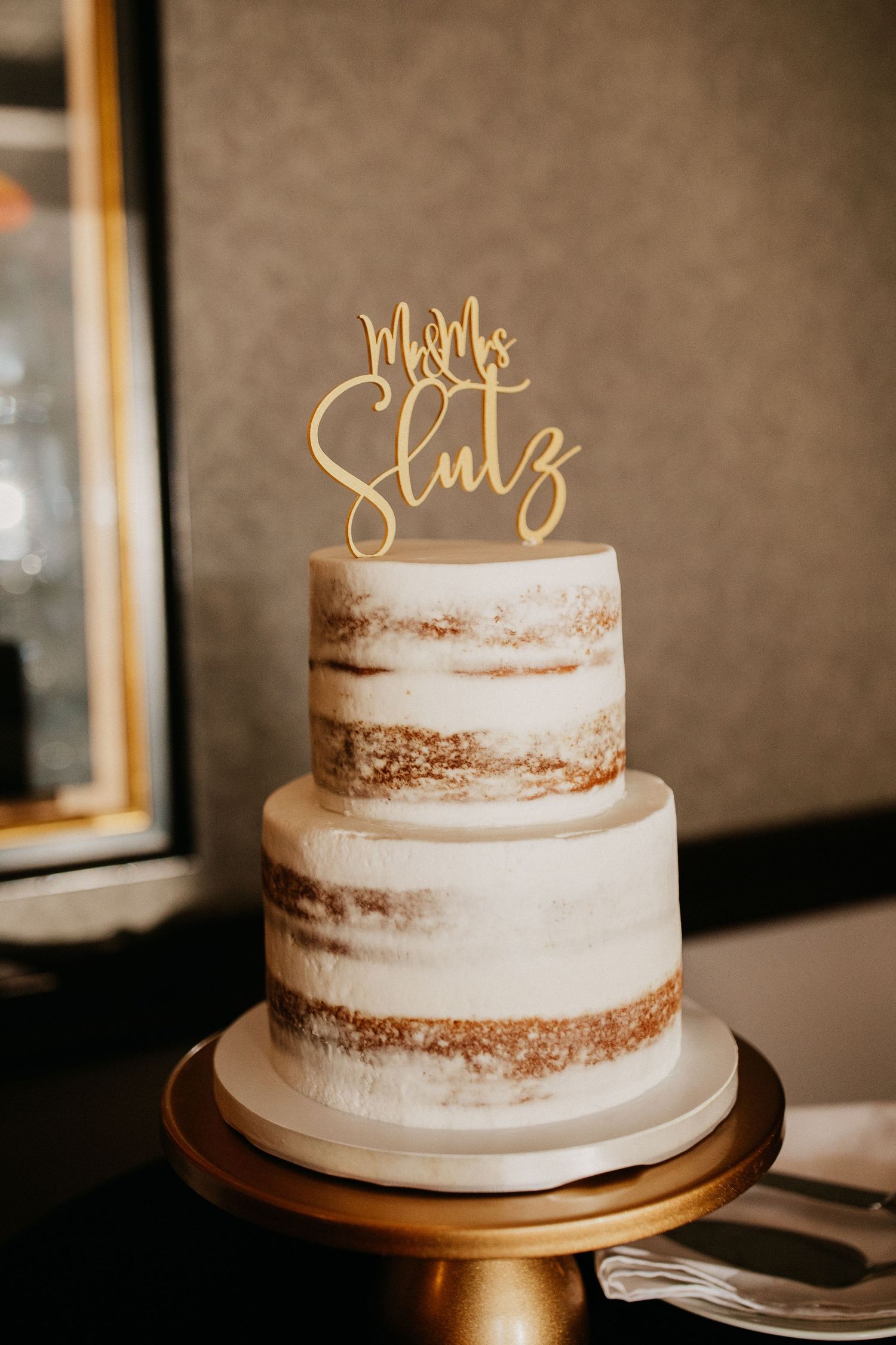 A two-tiered semi-naked wedding cake with a gold 'Mr & Mrs' topper on a gold stand.