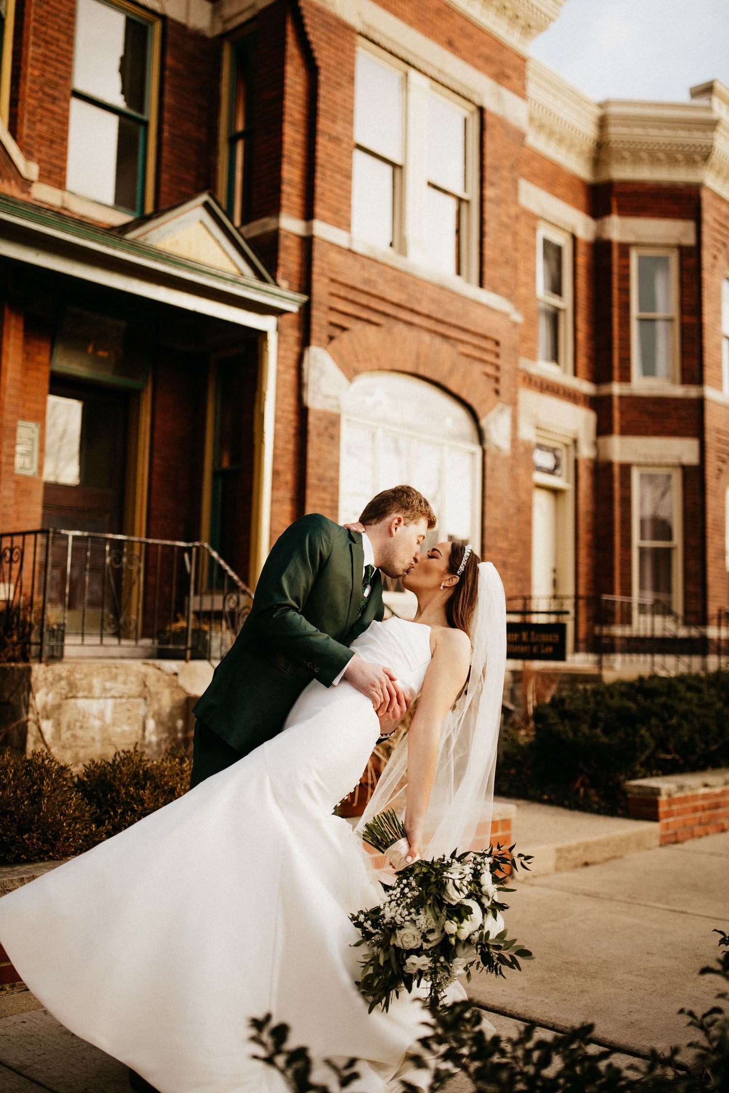 Elegant wedding couple poses against vintage brick architecture in warm sunlight.