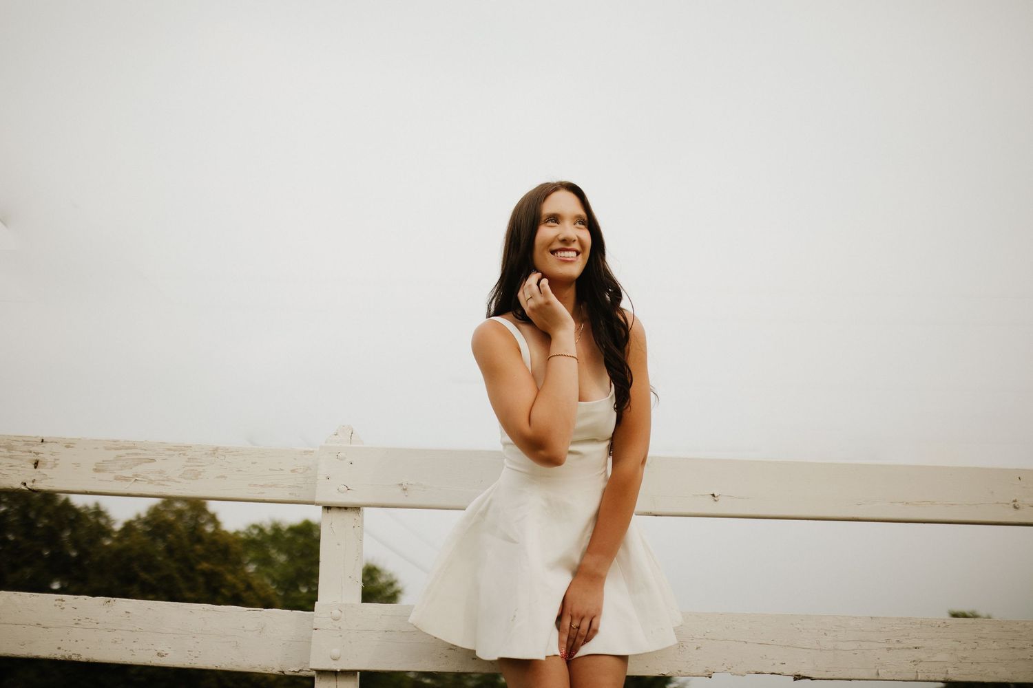 Person in white dress poses joyfully by a lake at sunset.