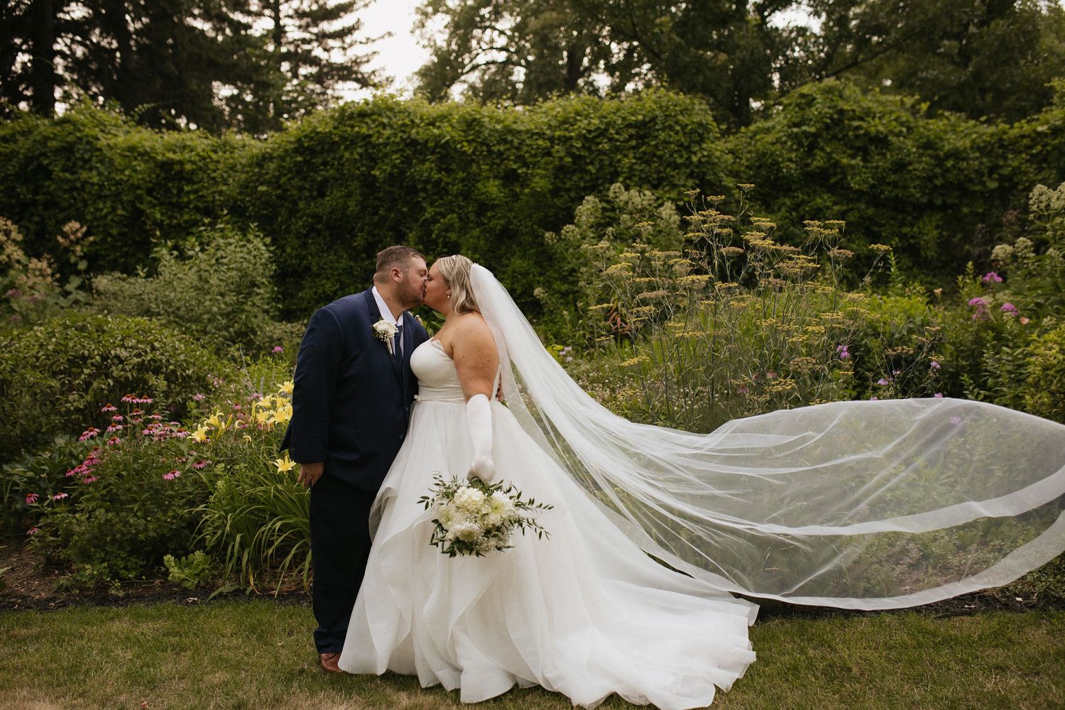 Wedding couple walks through garden setting with flowing veil trailing behind.