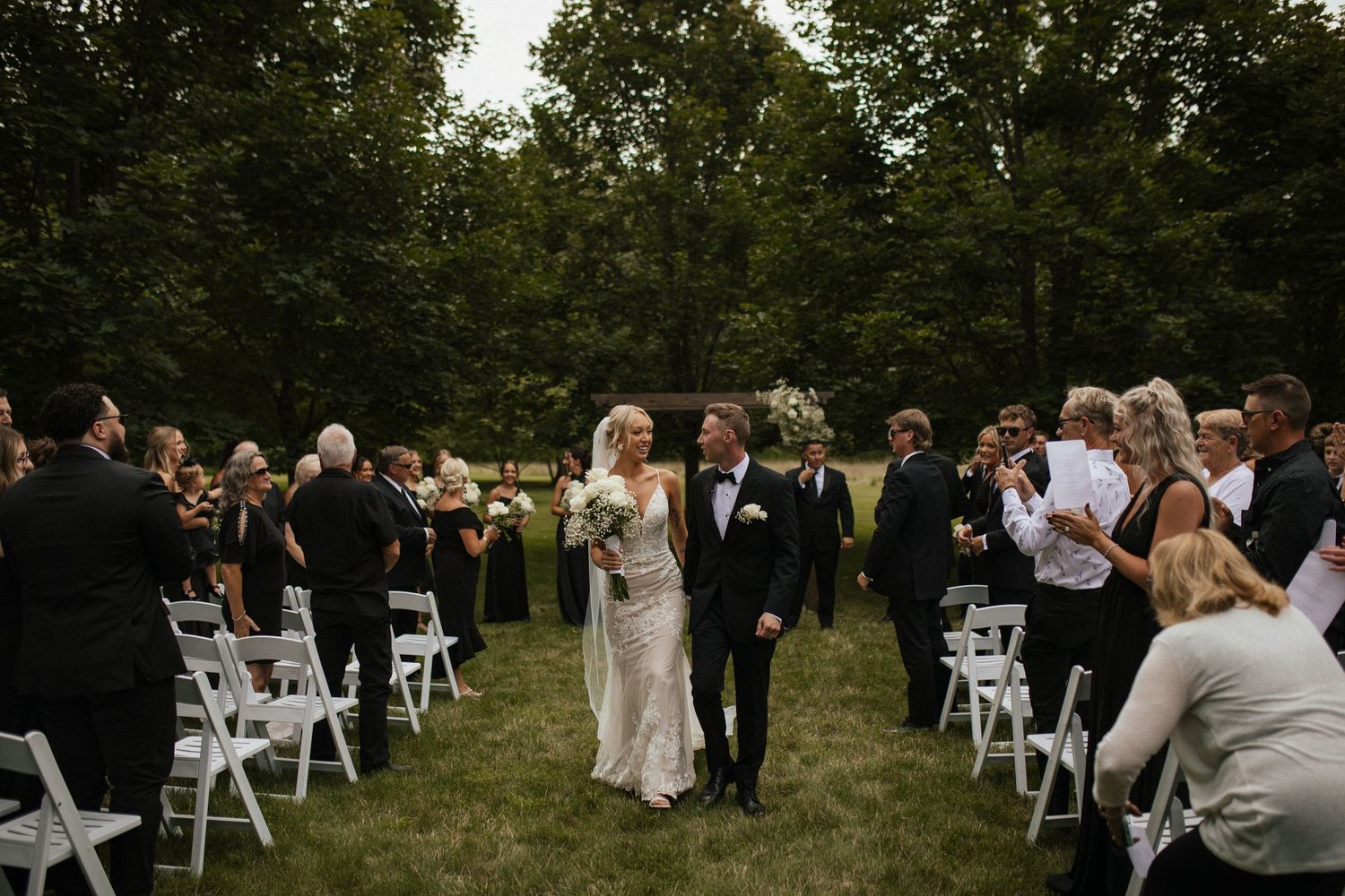 Outdoor wedding ceremony with rows of white chairs and guests gathered on a sunny day.