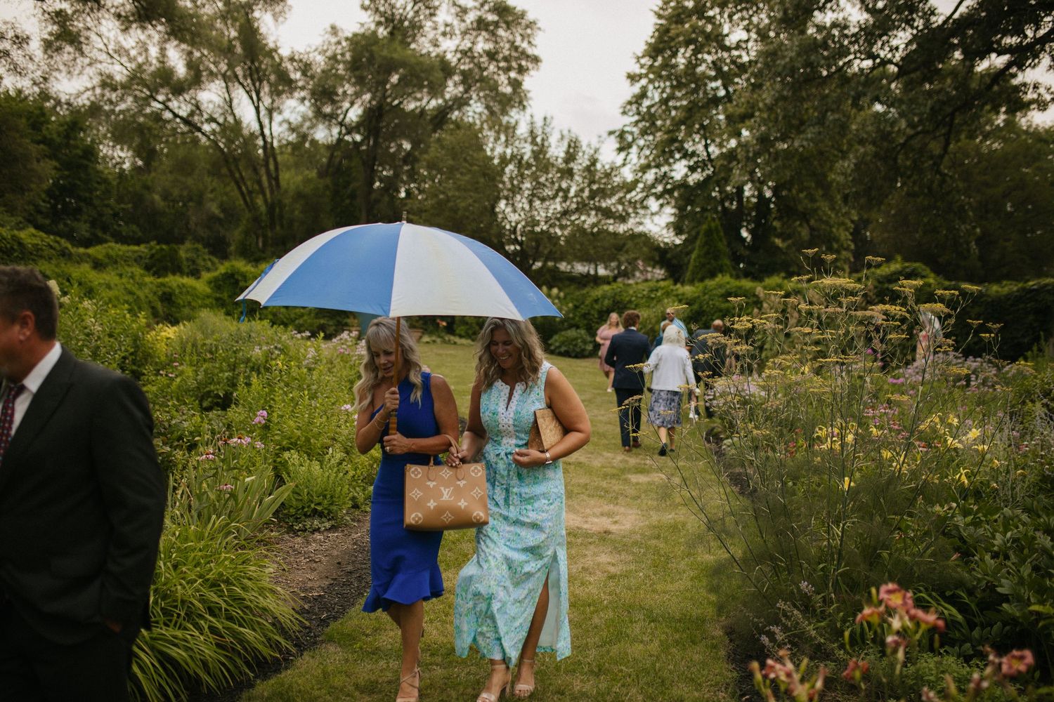 Wedding guests walk through a garden path carrying a blue umbrella on an overcast day.