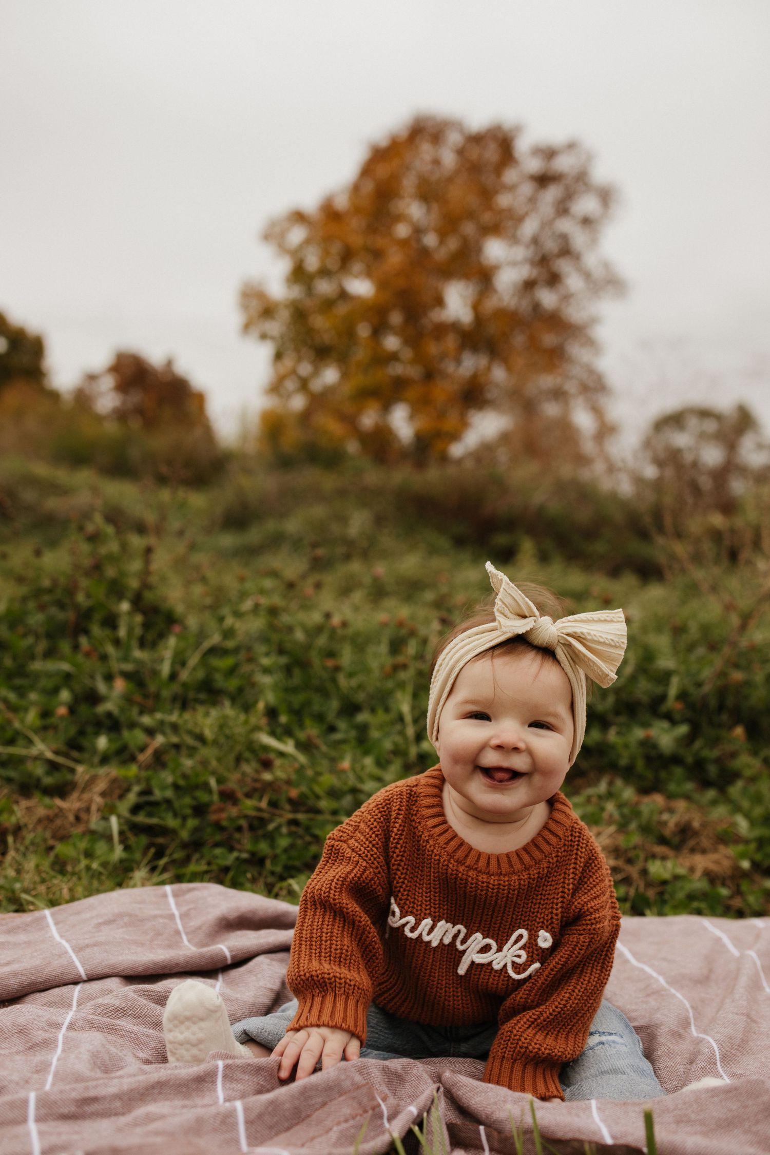 Smiling baby in rust-colored sweater and bow sits on blanket outdoors with fall foliage in background.