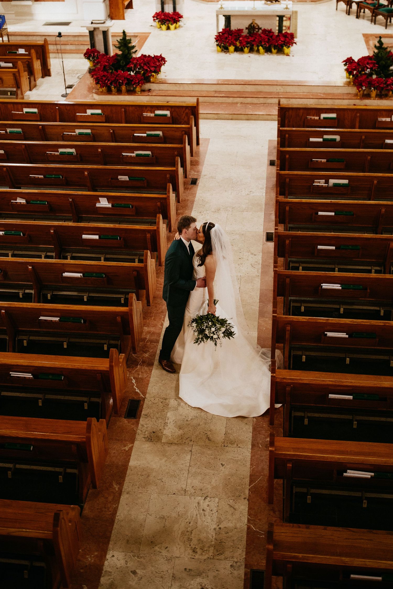 Aerial view of a couple in formal attire standing between wooden church pews decorated with holiday poinsettias.