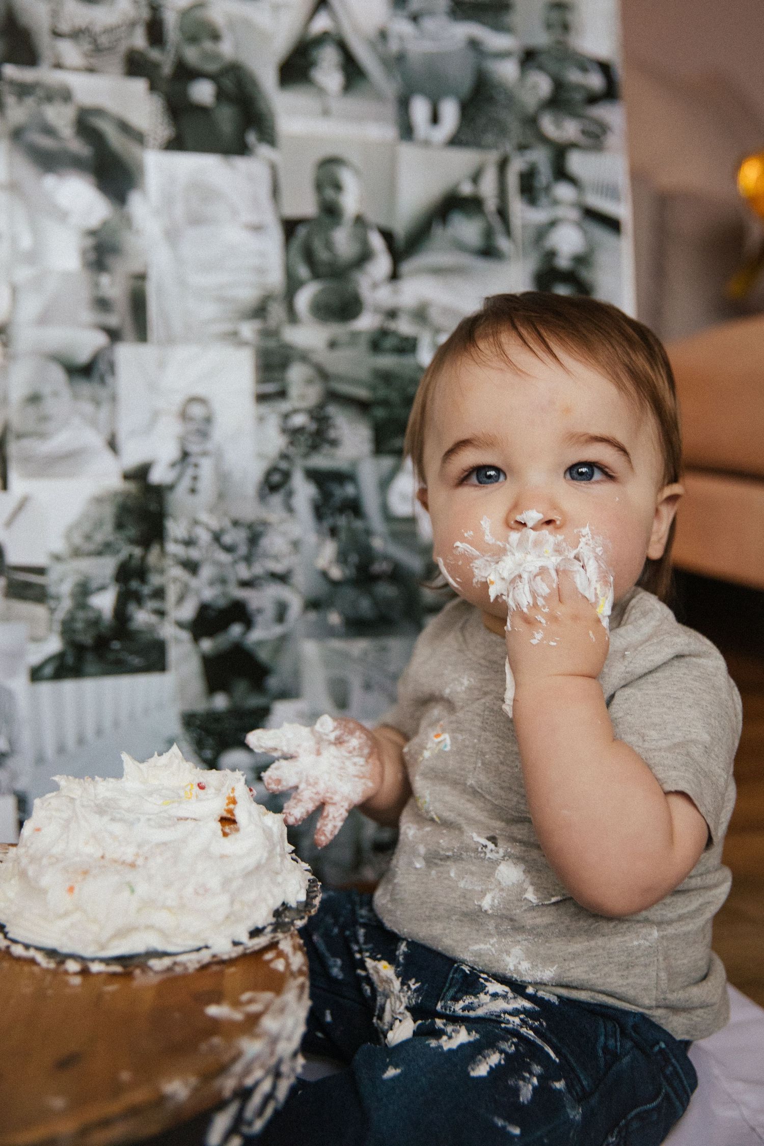 Toddler with cake frosting on face sits by black and white photo wall during birthday celebration.