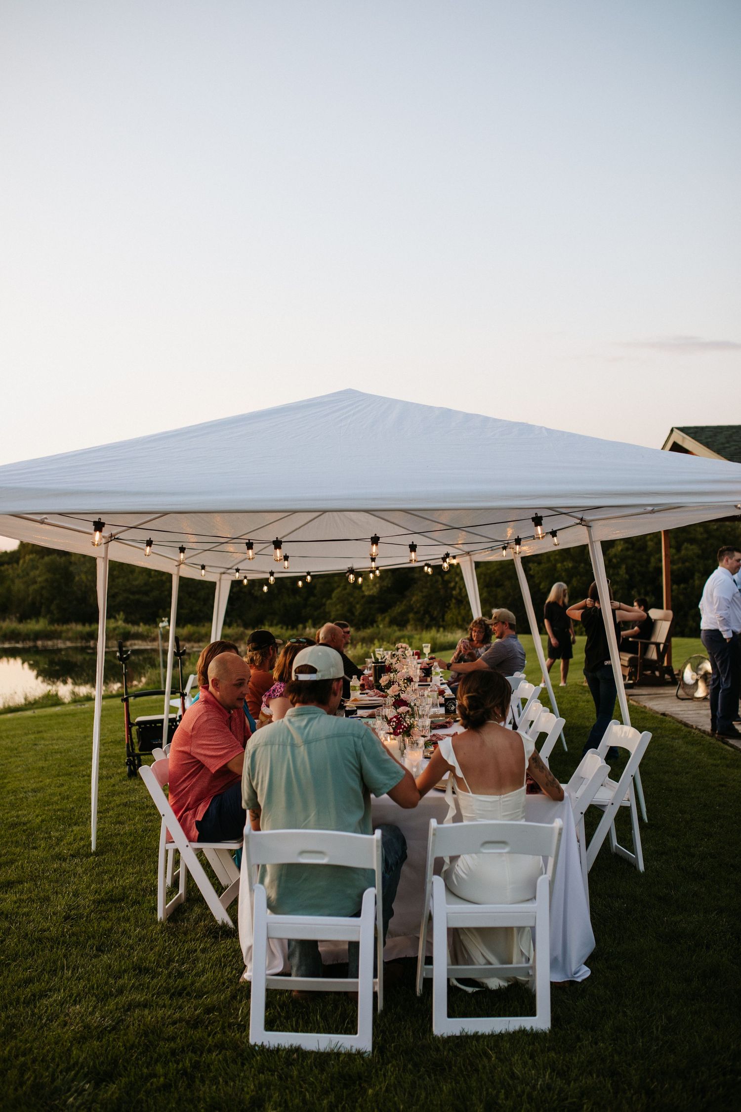 Outdoor wedding reception setup with white tents and chairs on a grassy field at sunset.