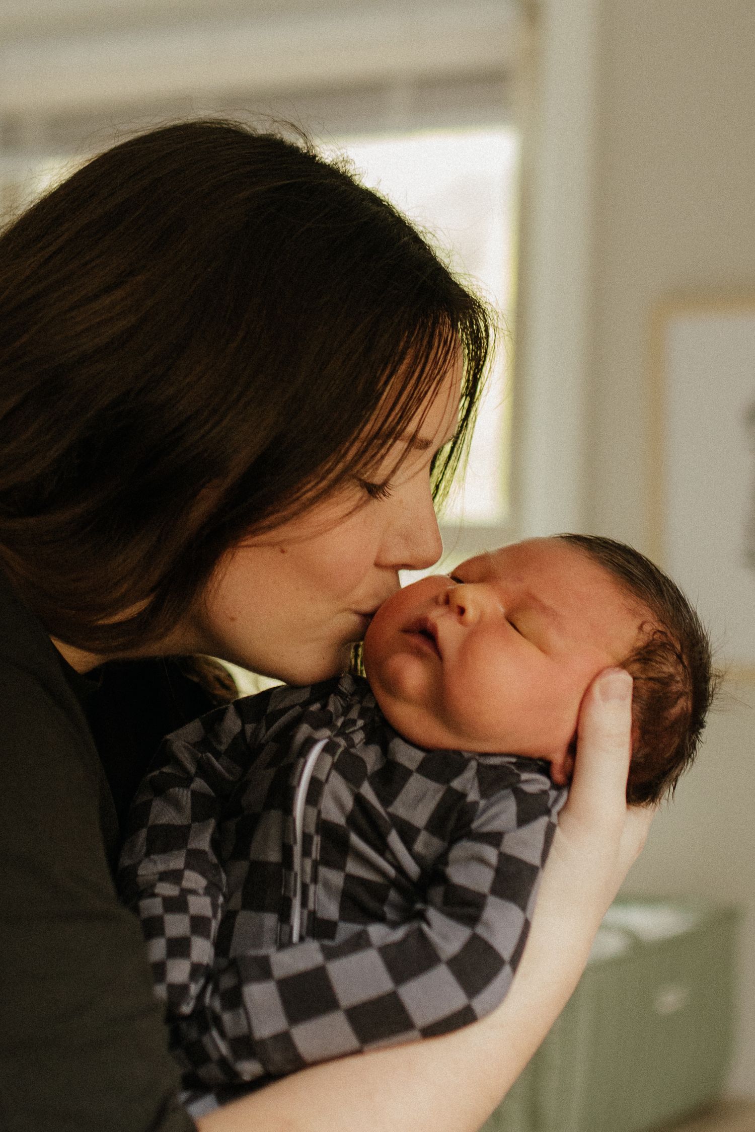 A mother tenderly kisses her baby wearing checkered clothing in soft natural light.