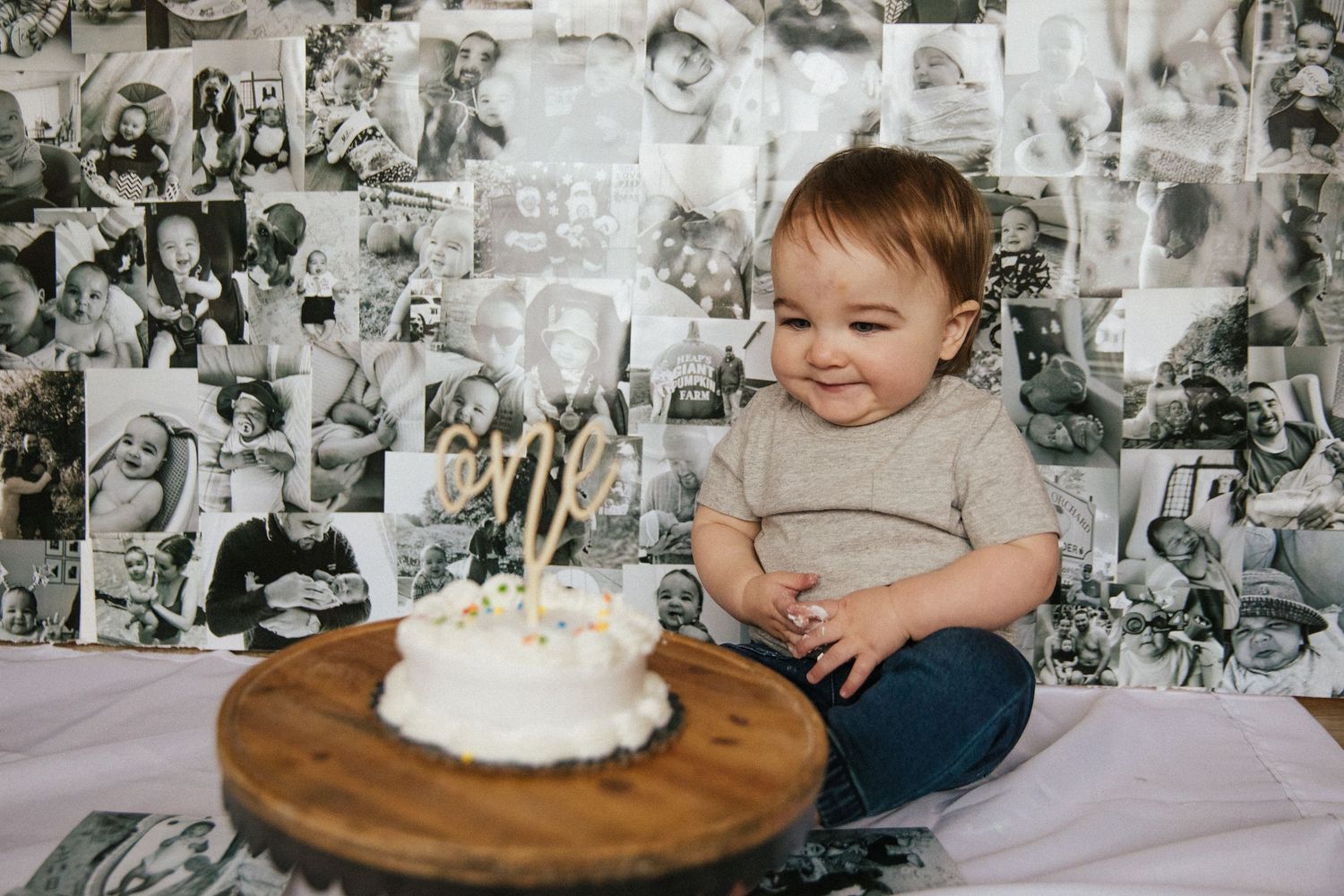 Smiling toddler sits next to birthday cake on wooden board against black and white photo wall.