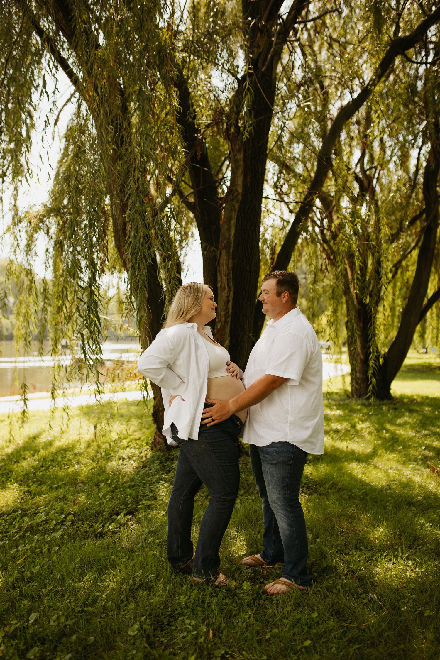 Couple standing together under weeping willow trees in sunlit park setting.