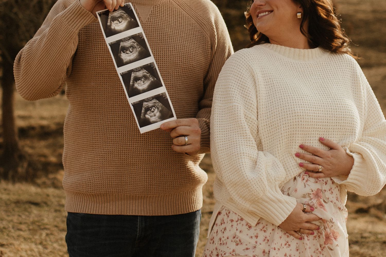 A couple holds ultrasound photos while embracing in warm sunlight.