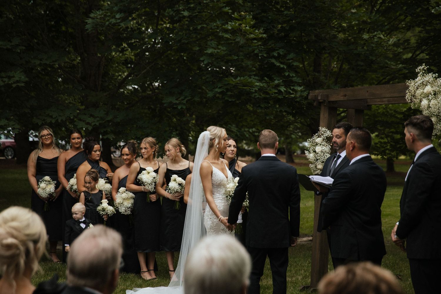 Wedding party and guests gathered for an intimate outdoor ceremony under trees.