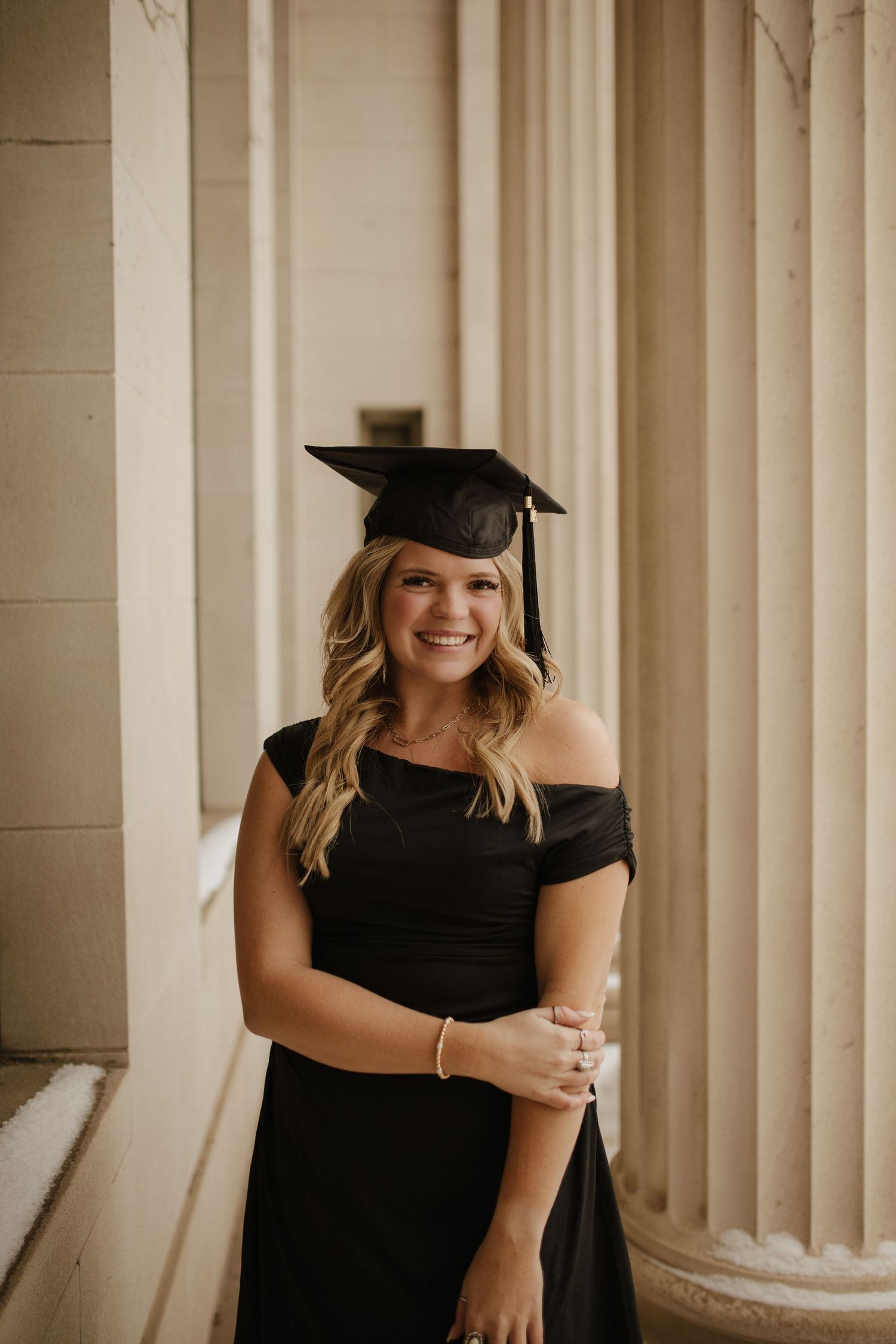 Graduate in black dress and cap posing by elegant columns.