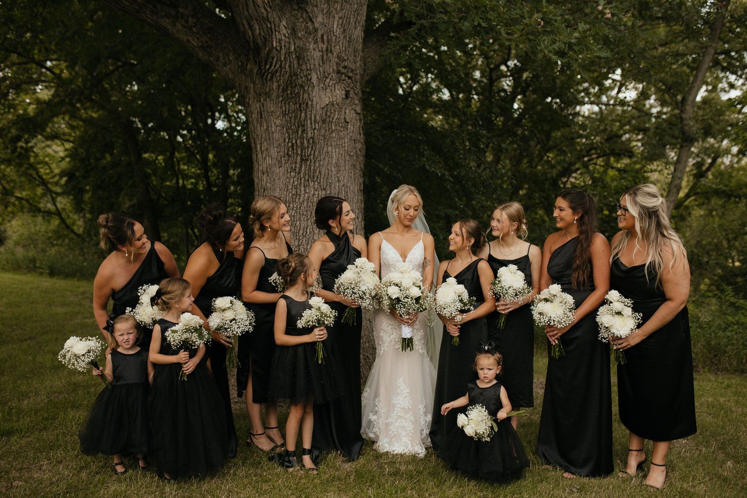 Wedding party in black dresses poses with bride against large tree trunk.