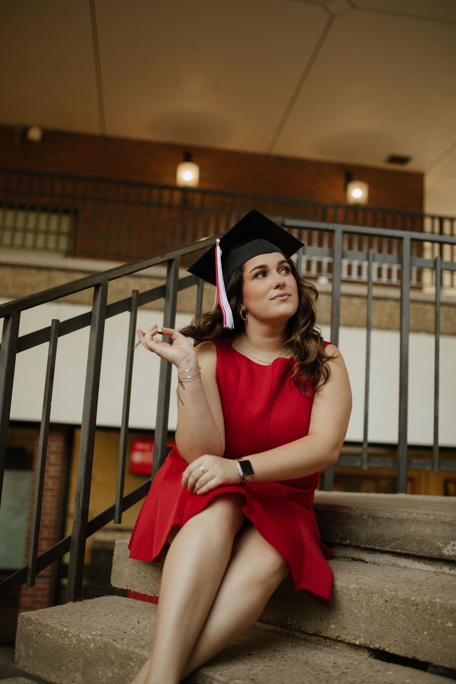 A graduate in red dress and black cap sits casually on concrete steps near metal railings.