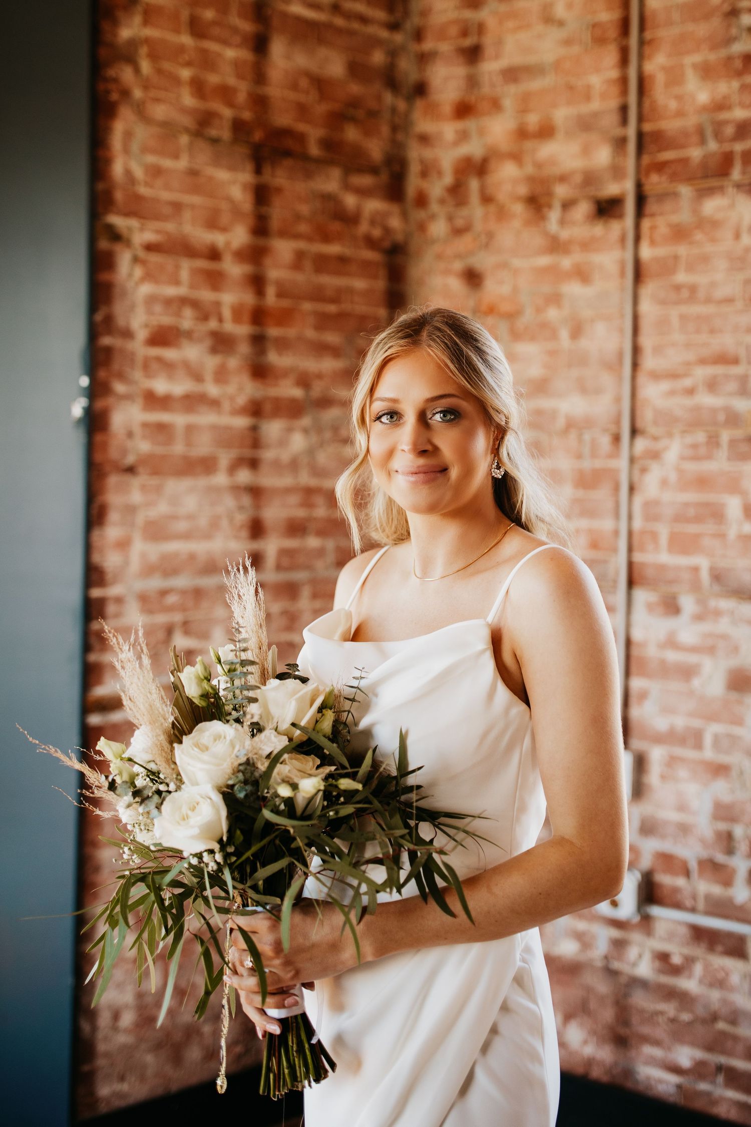 A bride holding a white and green bouquet poses against a rustic brick wall.