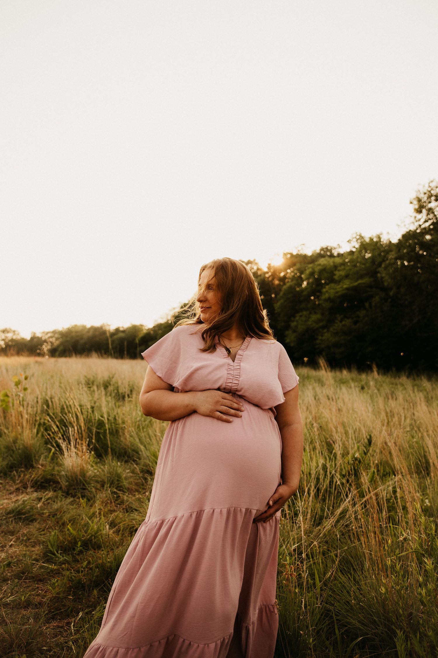 Expectant mother in pink maxi dress stands in golden field at sunset.
