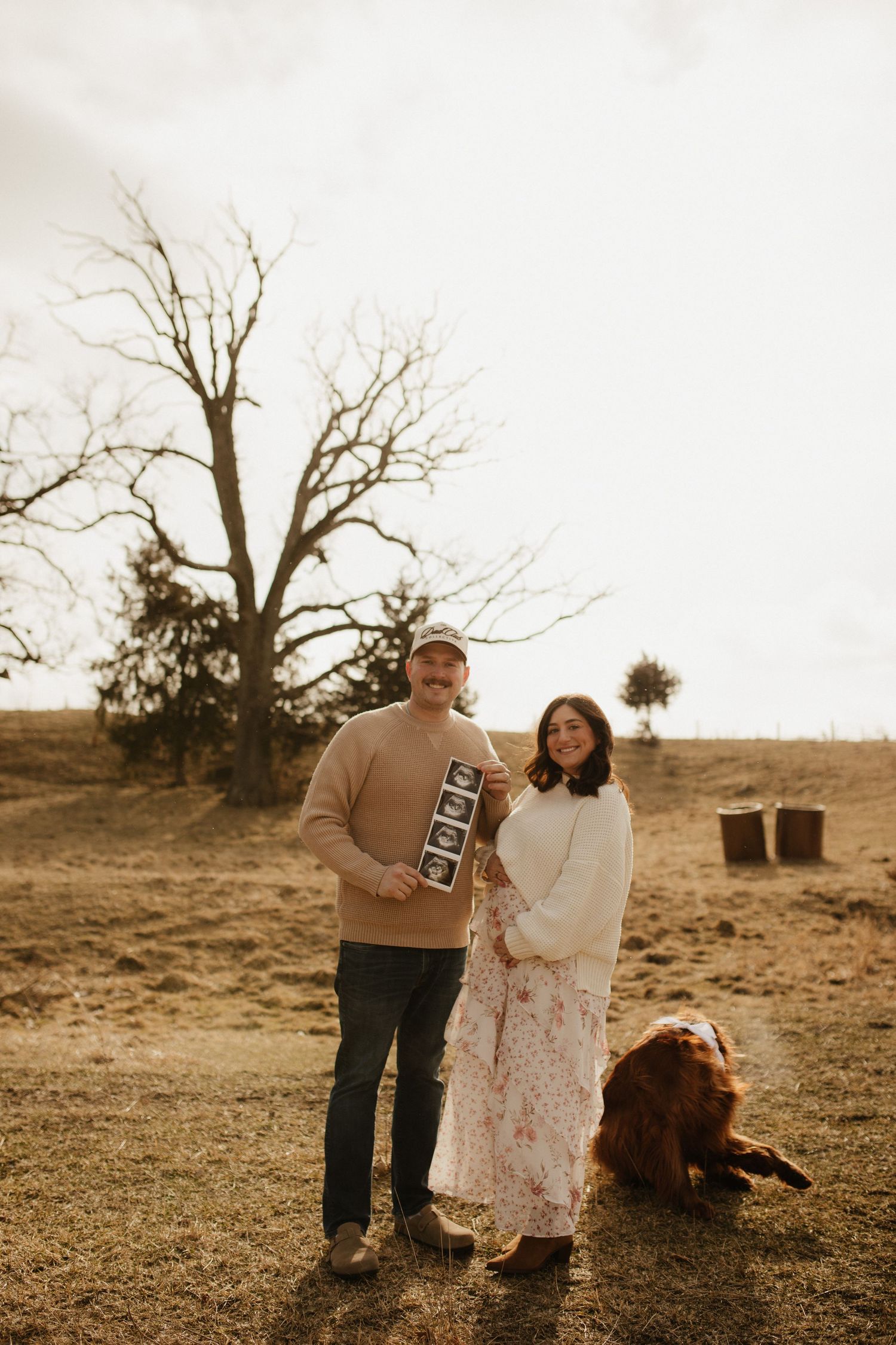A couple and their dog stand together in a sepia-toned outdoor setting holding ultrasound photos.