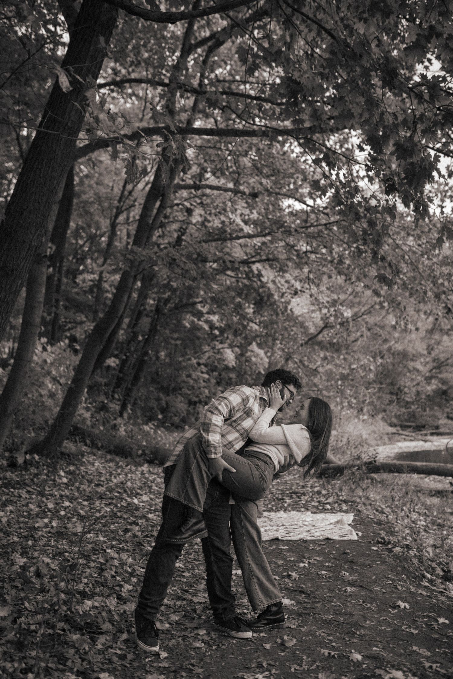 Romantic black and white photo of a couple kissing on a leaf-covered forest path.
