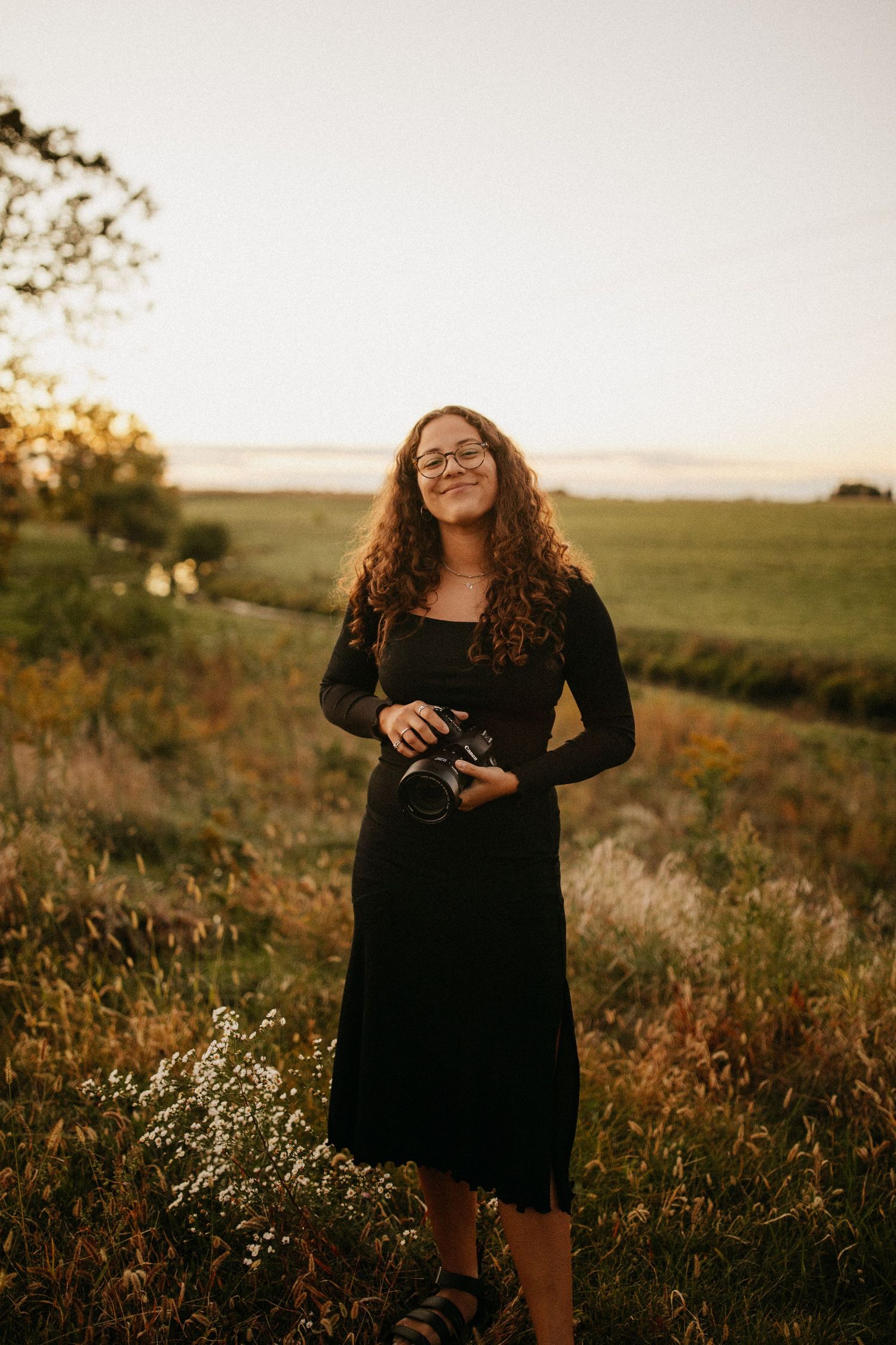 Photographer in a black dress stands in a golden field at sunset holding a camera.