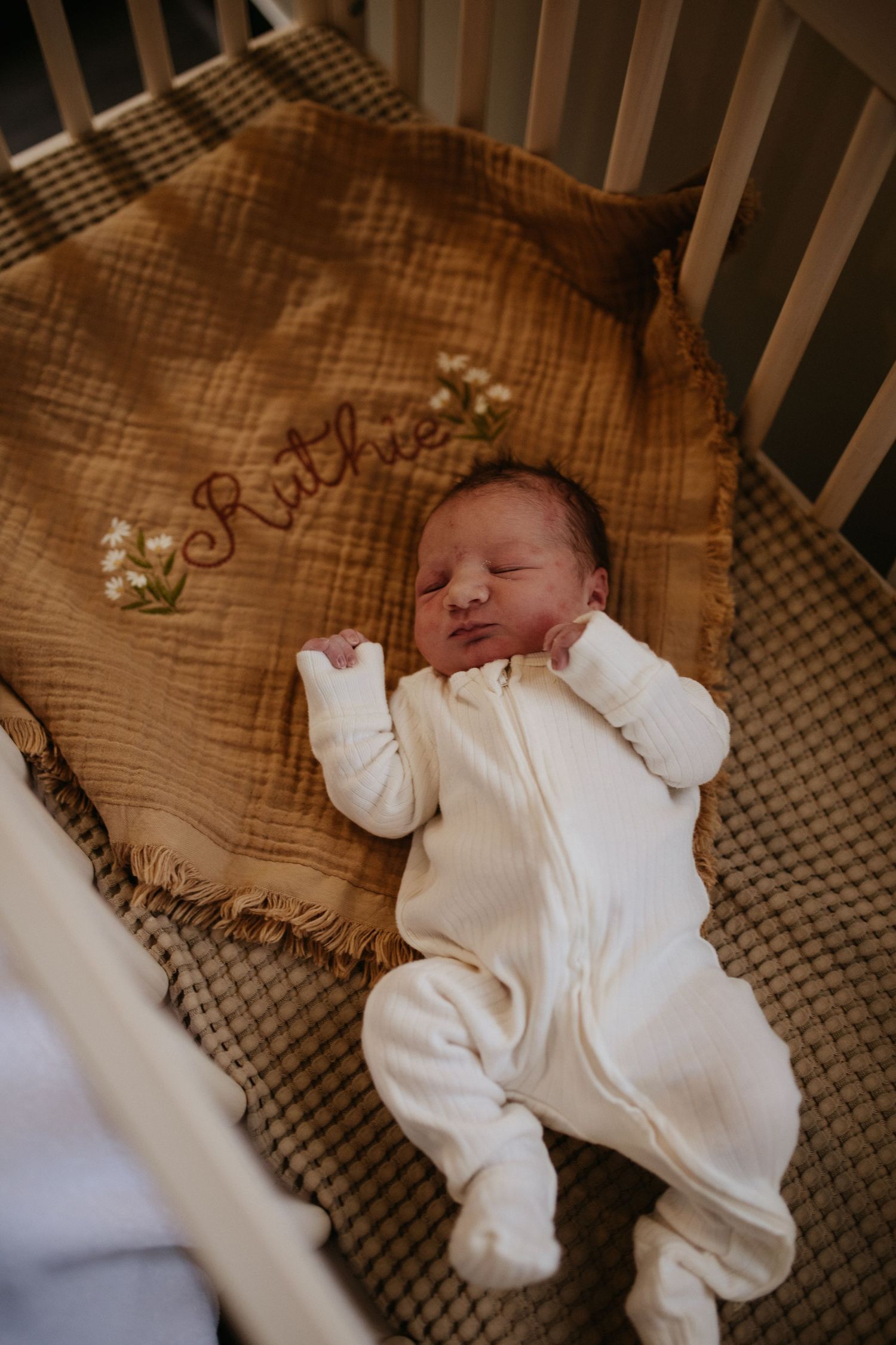 Baby sleeping in white onesie on brown embroidered crib blanket.