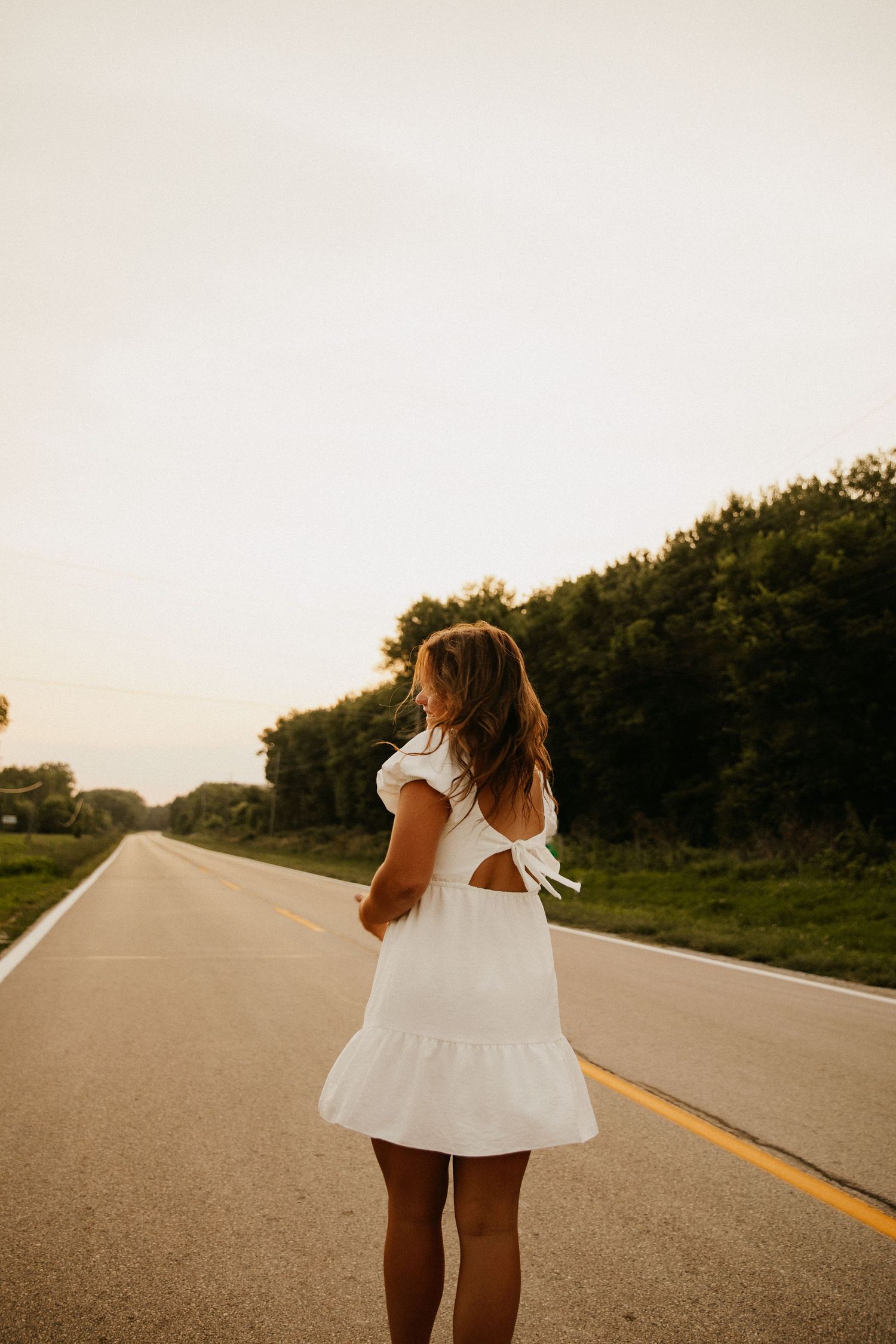 Person in white summer dress walks along empty rural road at sunset.