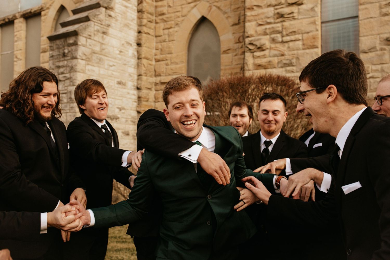 Groomsmen in black suits sharing a lighthearted moment outside a stone church.