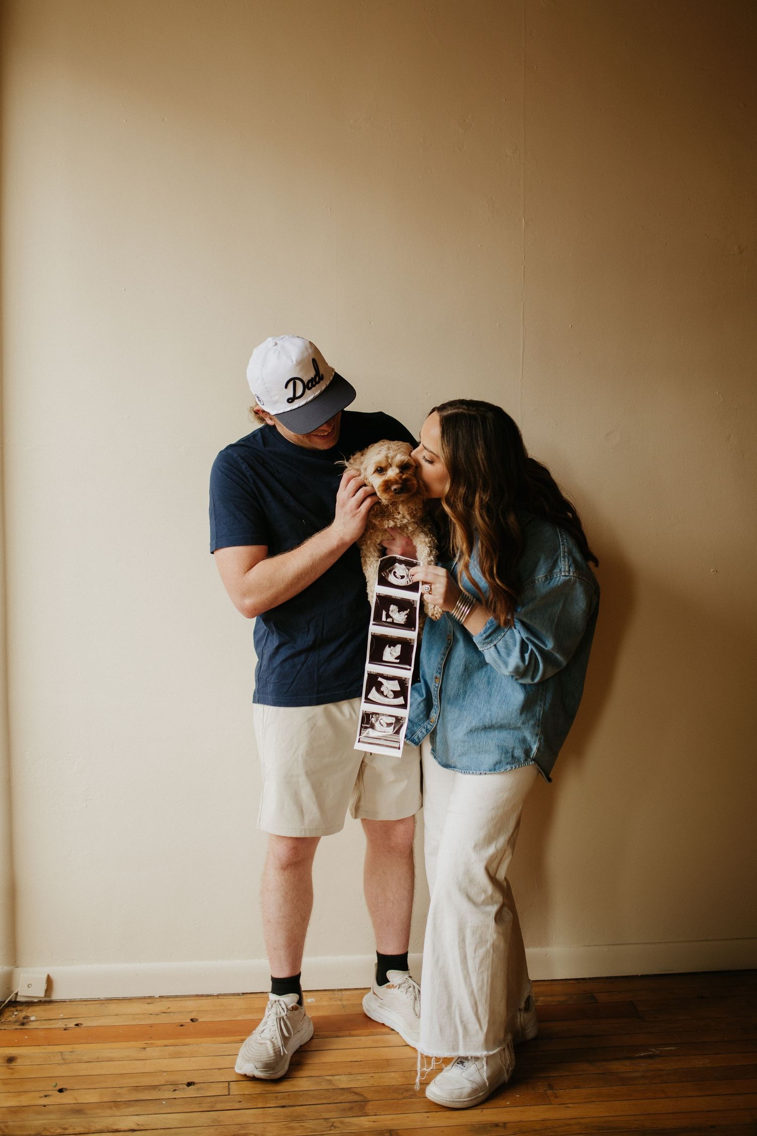 A couple sits on wooden floors holding ultrasound photos in a playful pose.