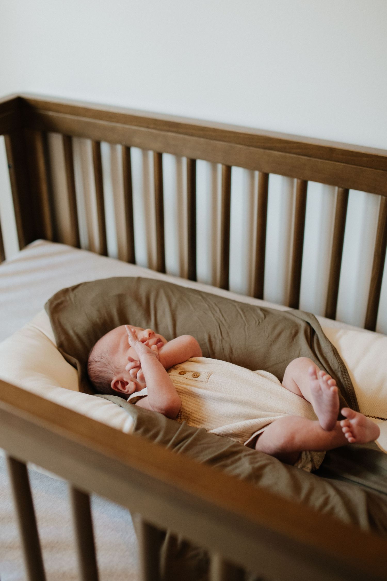 Sleeping newborn stretches in wooden crib with neutral bedding.