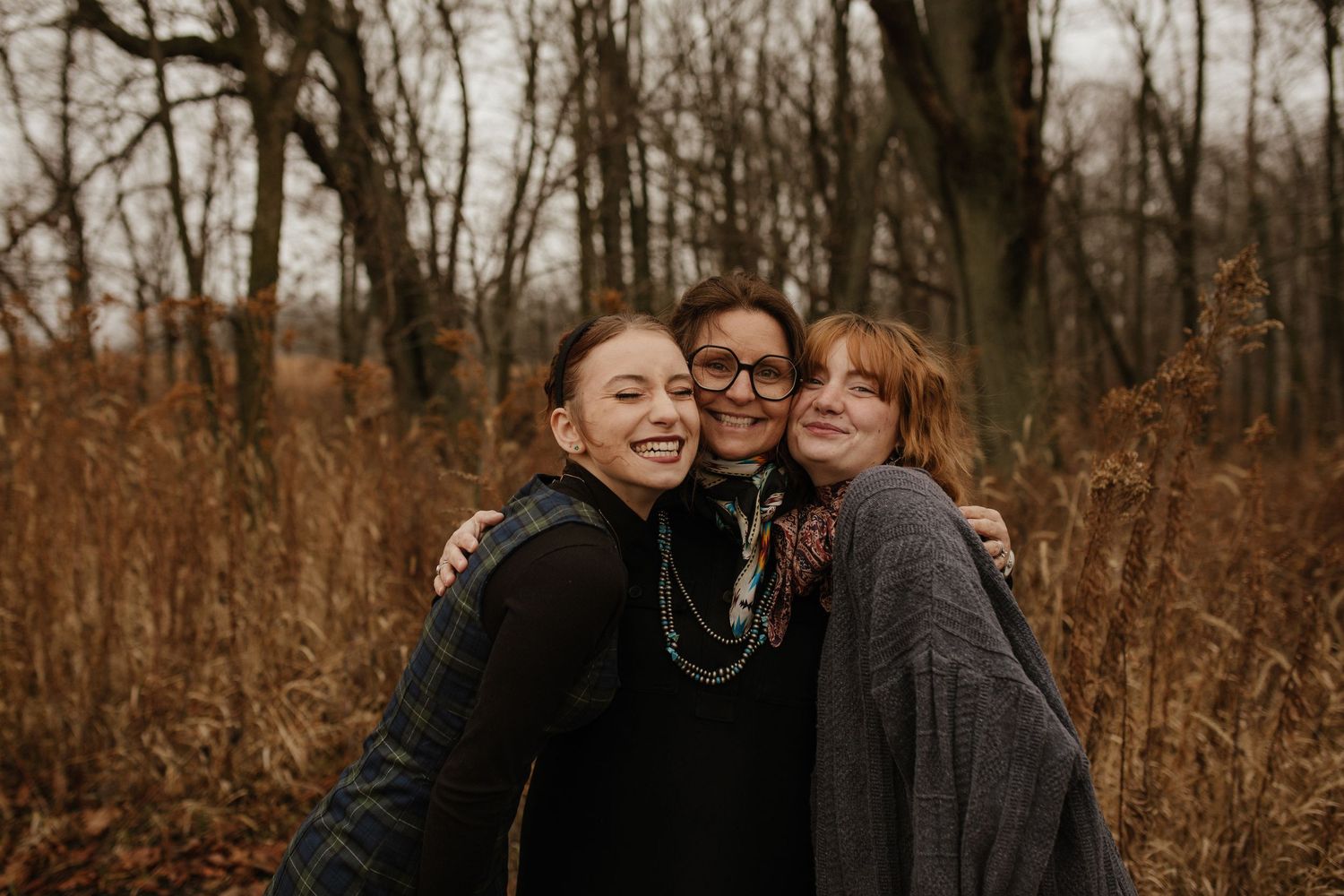 Three friends share a joyful embrace while standing in tall autumn grass with bare trees in the background.