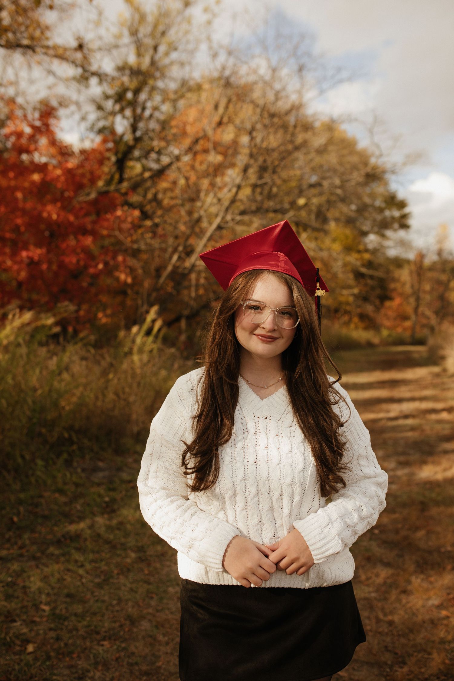 A person wearing a burgundy beret and white cable knit sweater poses outdoors in fall scenery.