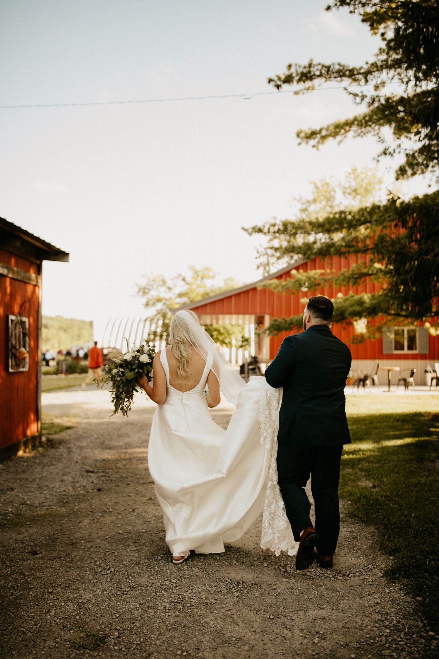 Couple walks along path near red barn buildings during sunset wedding photos.