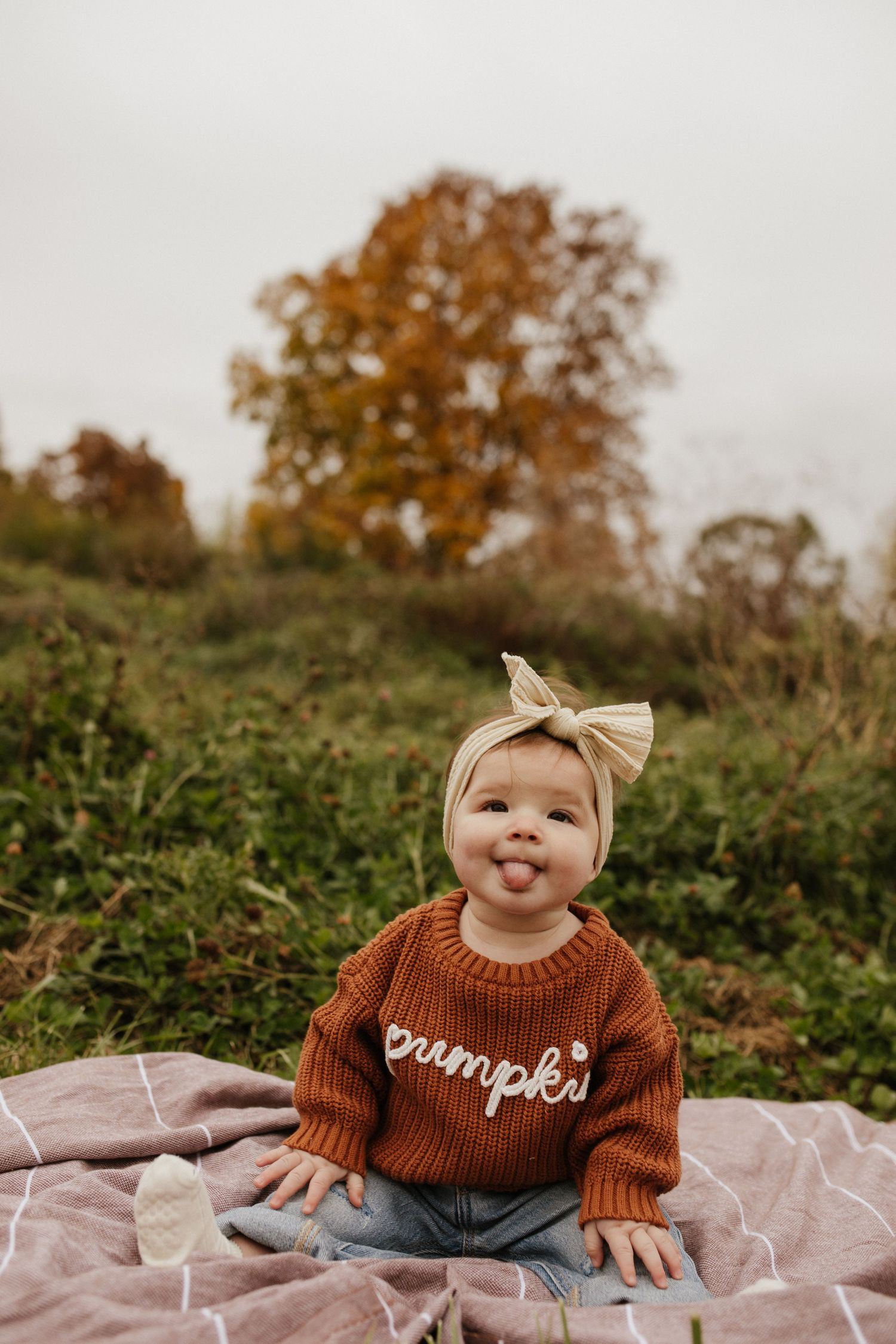 Baby wearing brown sweater with 'pumpkin' text and bow headband sits on blanket during autumn photoshoot.