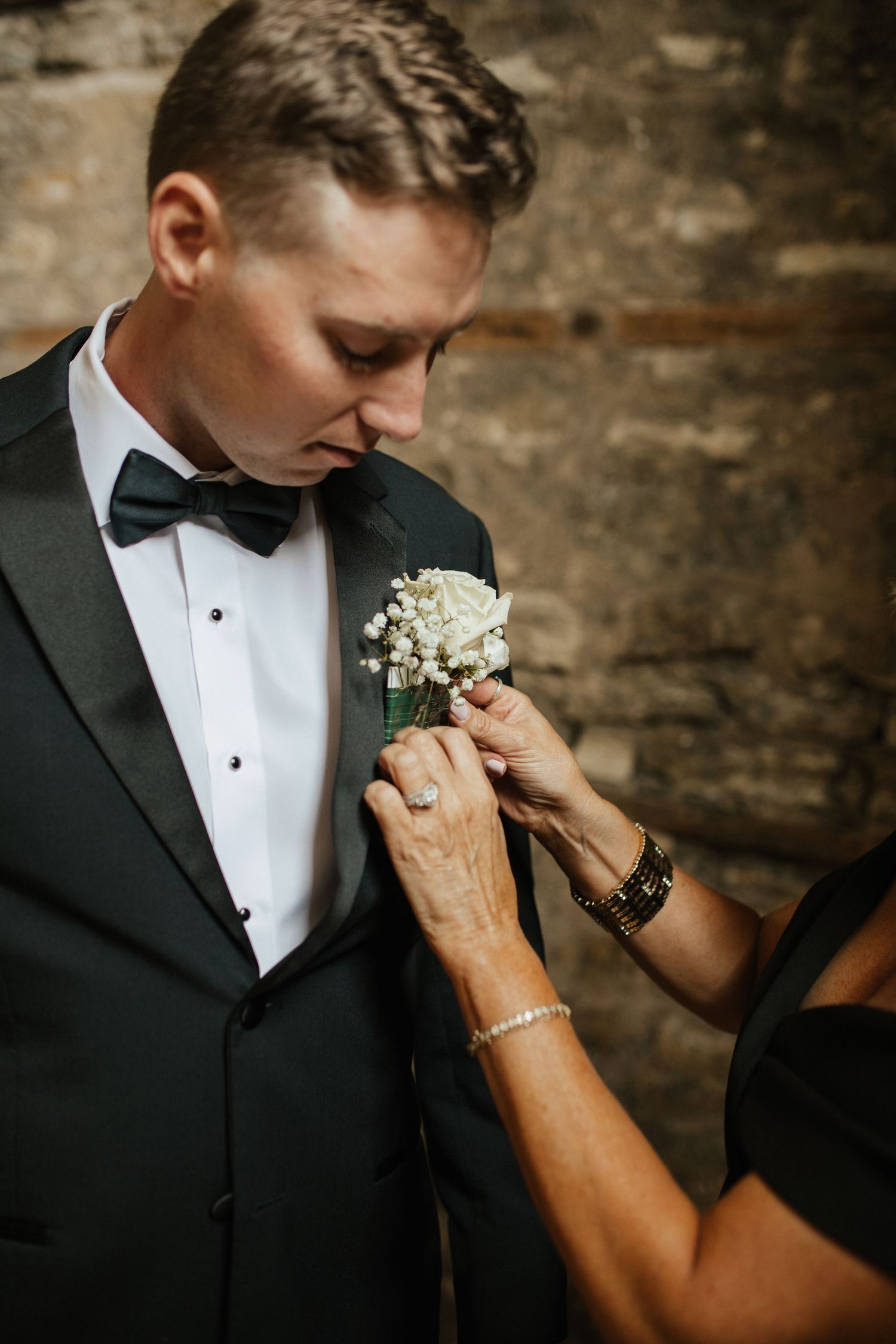 Close-up shots of hands pinning a white boutonniere on a groom's black tuxedo lapel.