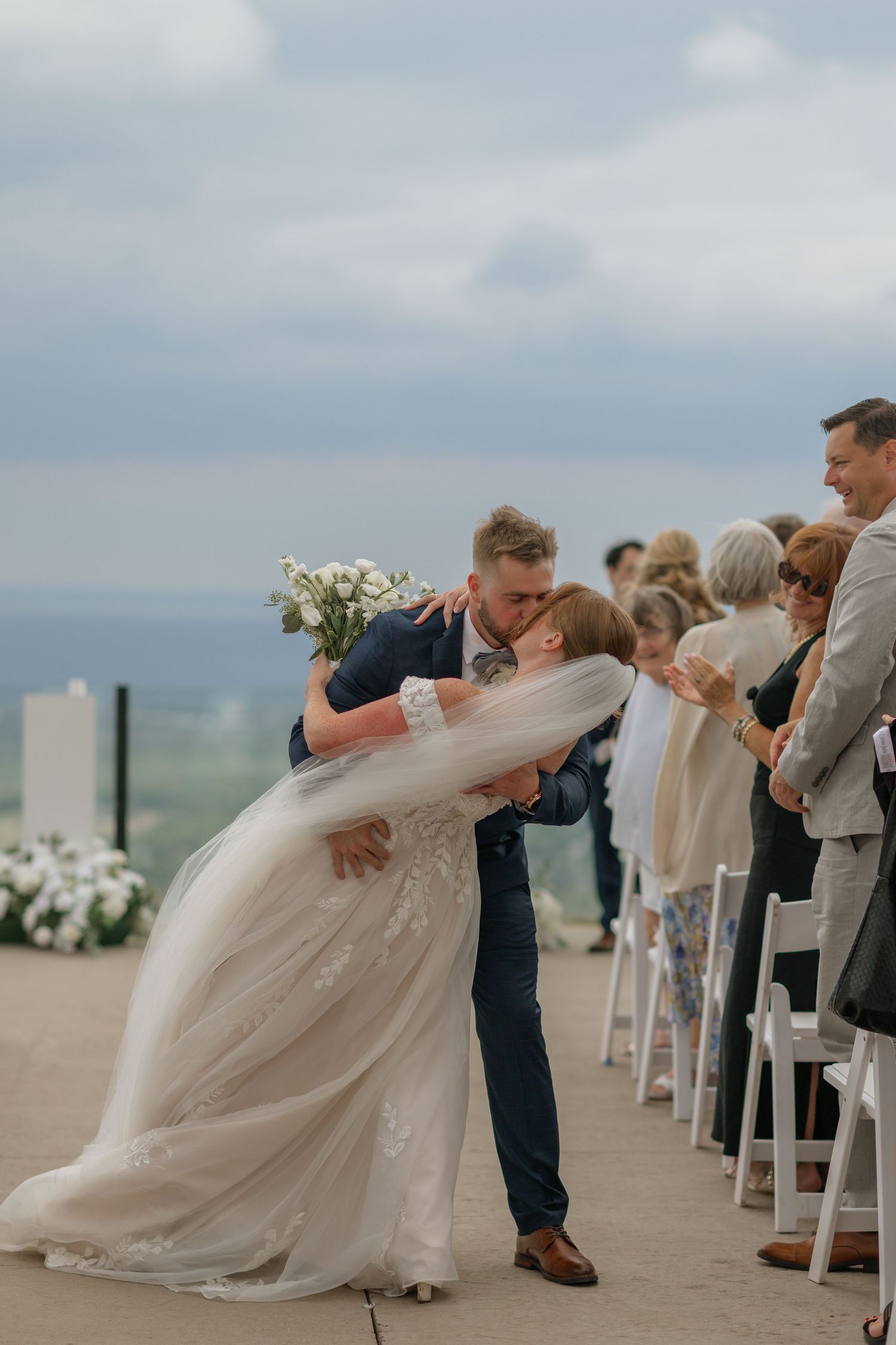 A romantic first kiss during an outdoor wedding ceremony with guests looking on against a cloudy sky.