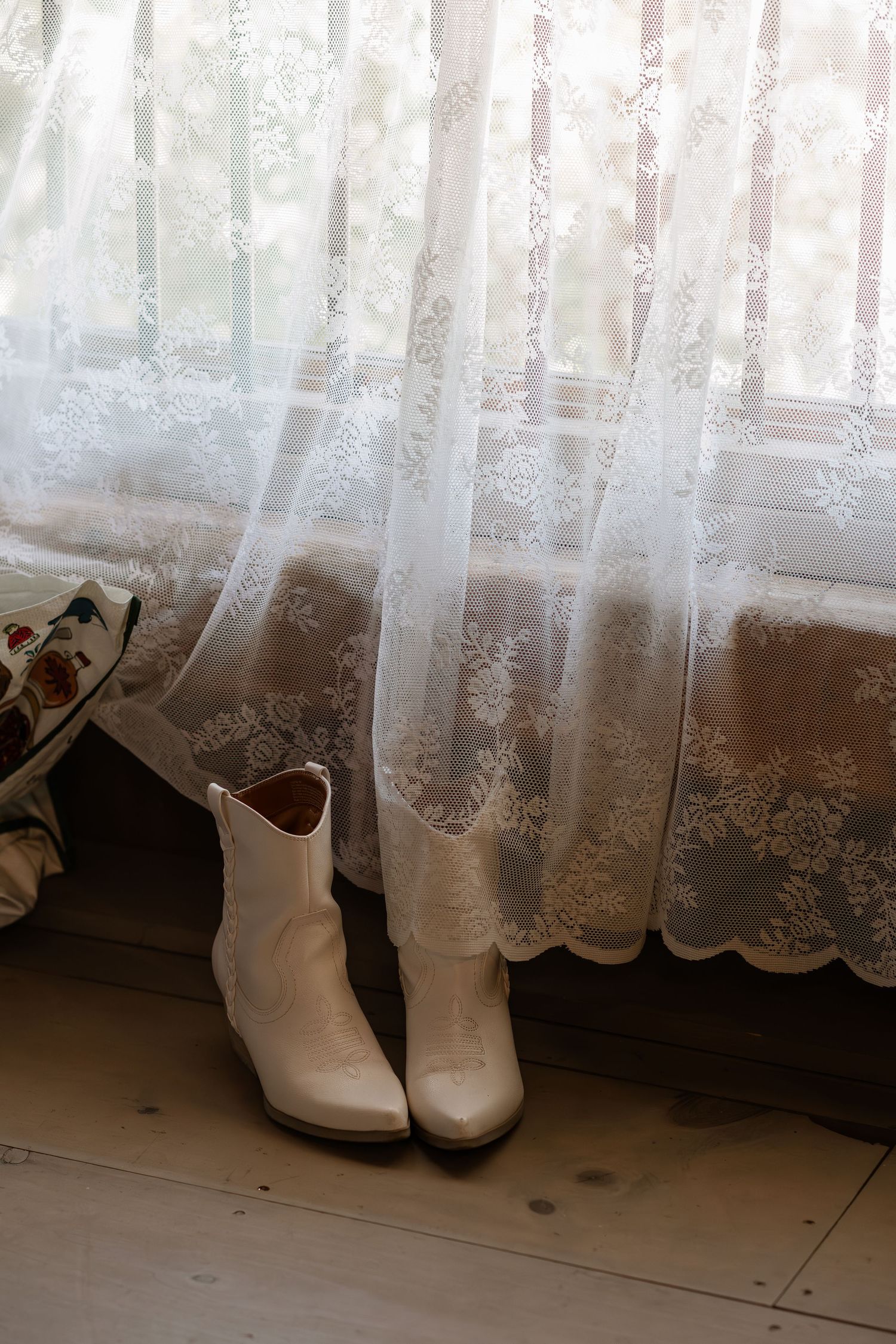 White lace curtains and cowboy boots line up along a rustic wooden floor near a window.