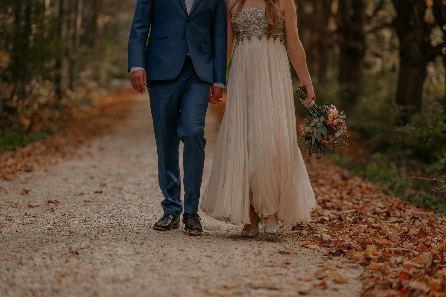 A couple walks through fallen autumn leaves on a forest path in formal wedding attire.